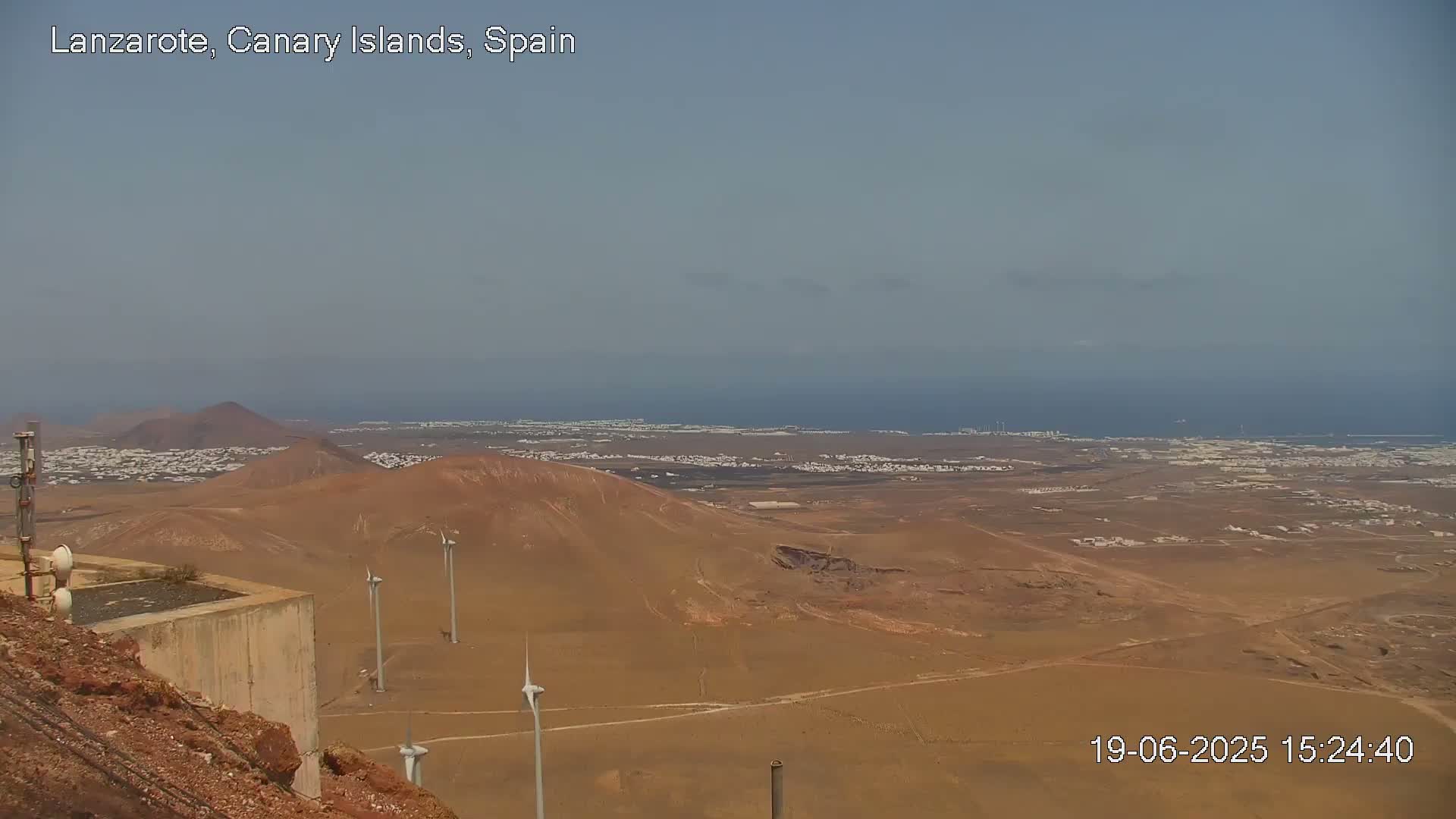 A high-angle, long shot shows a desert landscape with wind turbines, a town in the distance, and the ocean under a hazy sky.
