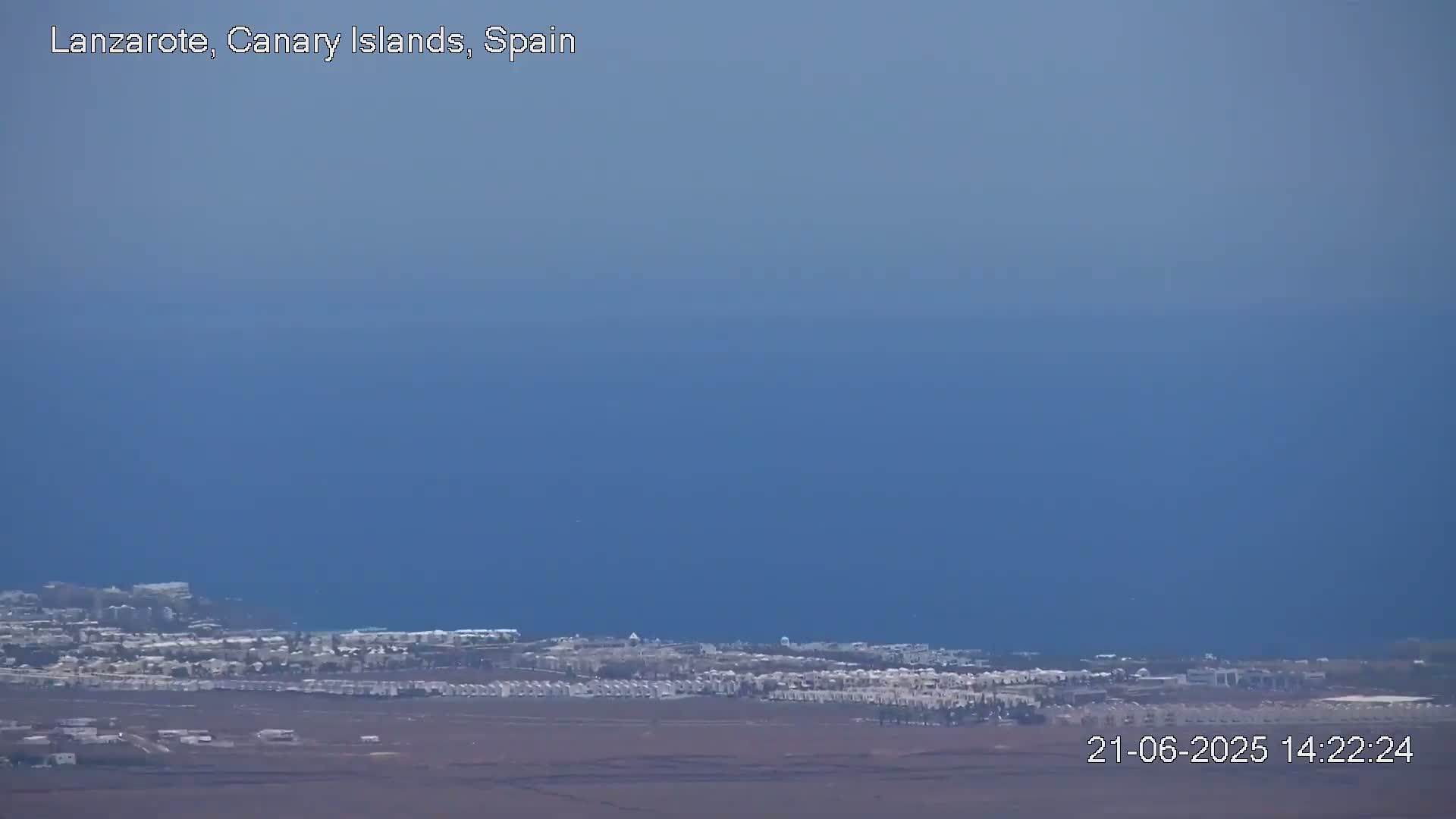 A coastal town with low-rise buildings stretches along the ocean under a clear, pale blue sky.