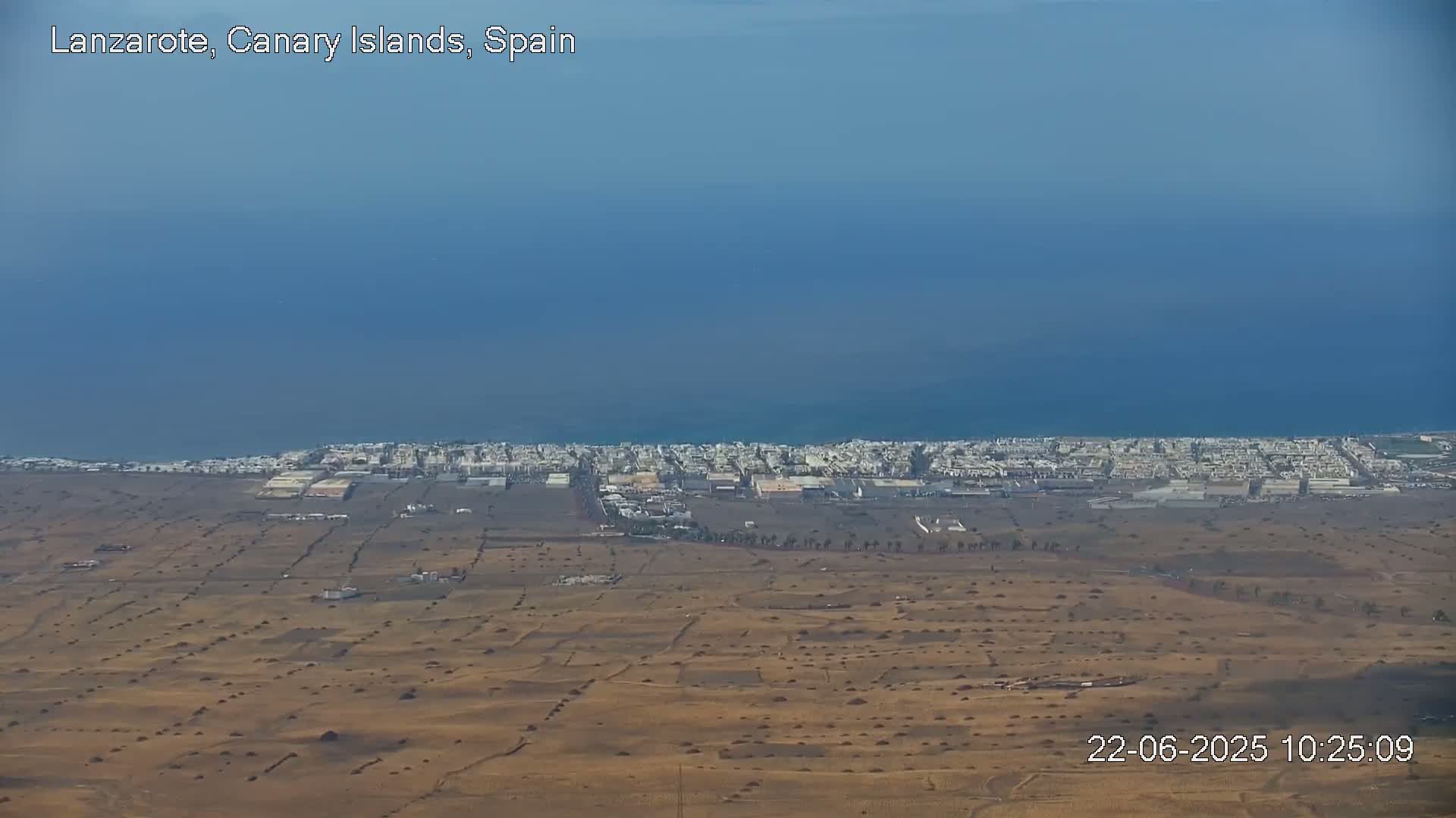 A coastal town with light-colored buildings is visible beside a calm blue ocean under a clear sky, with a large expanse of dry, brown land stretching towards the viewer.