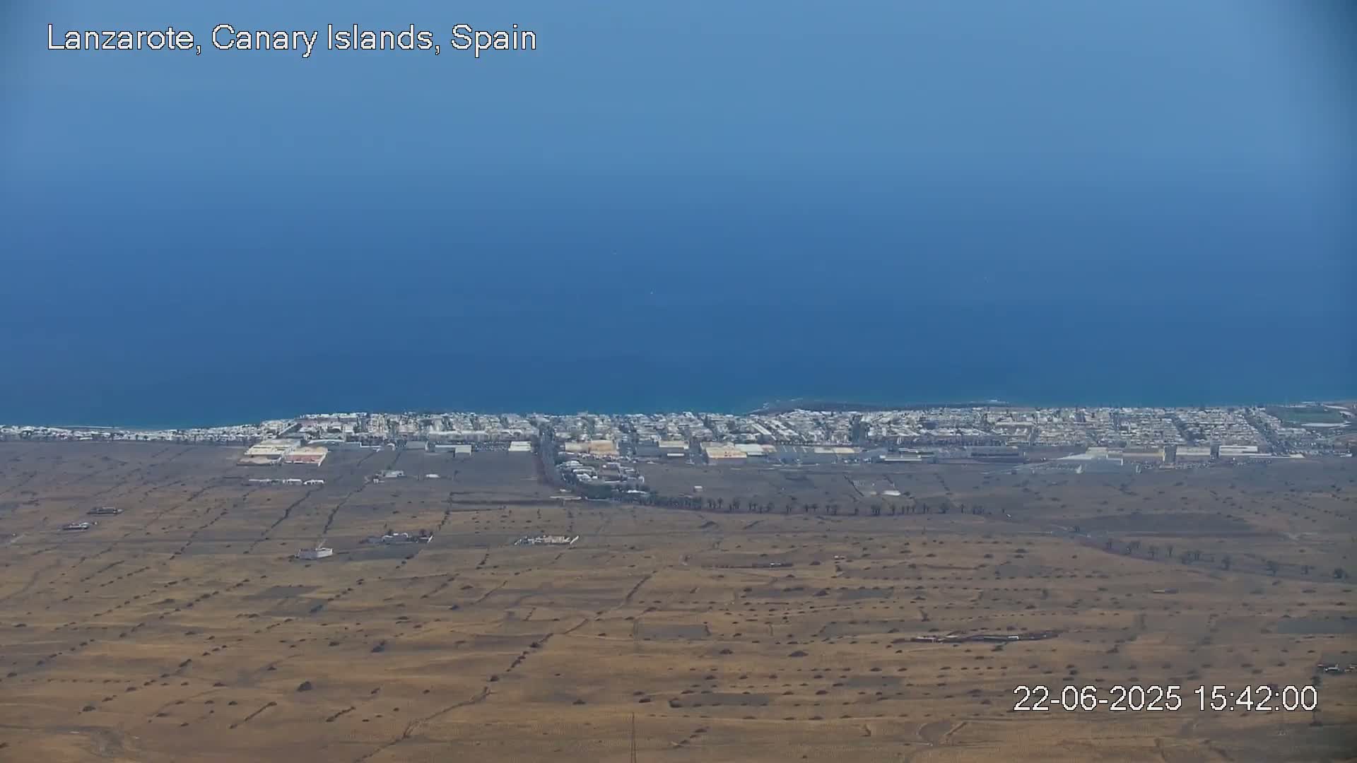 A coastal town with a dry, brown landscape stretches towards a calm blue ocean under a clear sky.