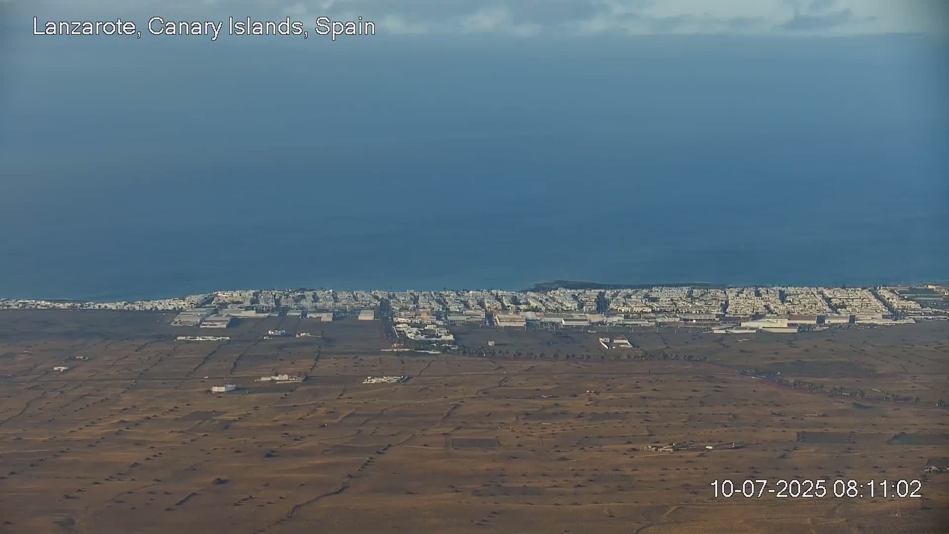 A coastal town with light-colored buildings stretches along a dark-brown, arid landscape next to a calm, blue ocean under a mostly clear sky.