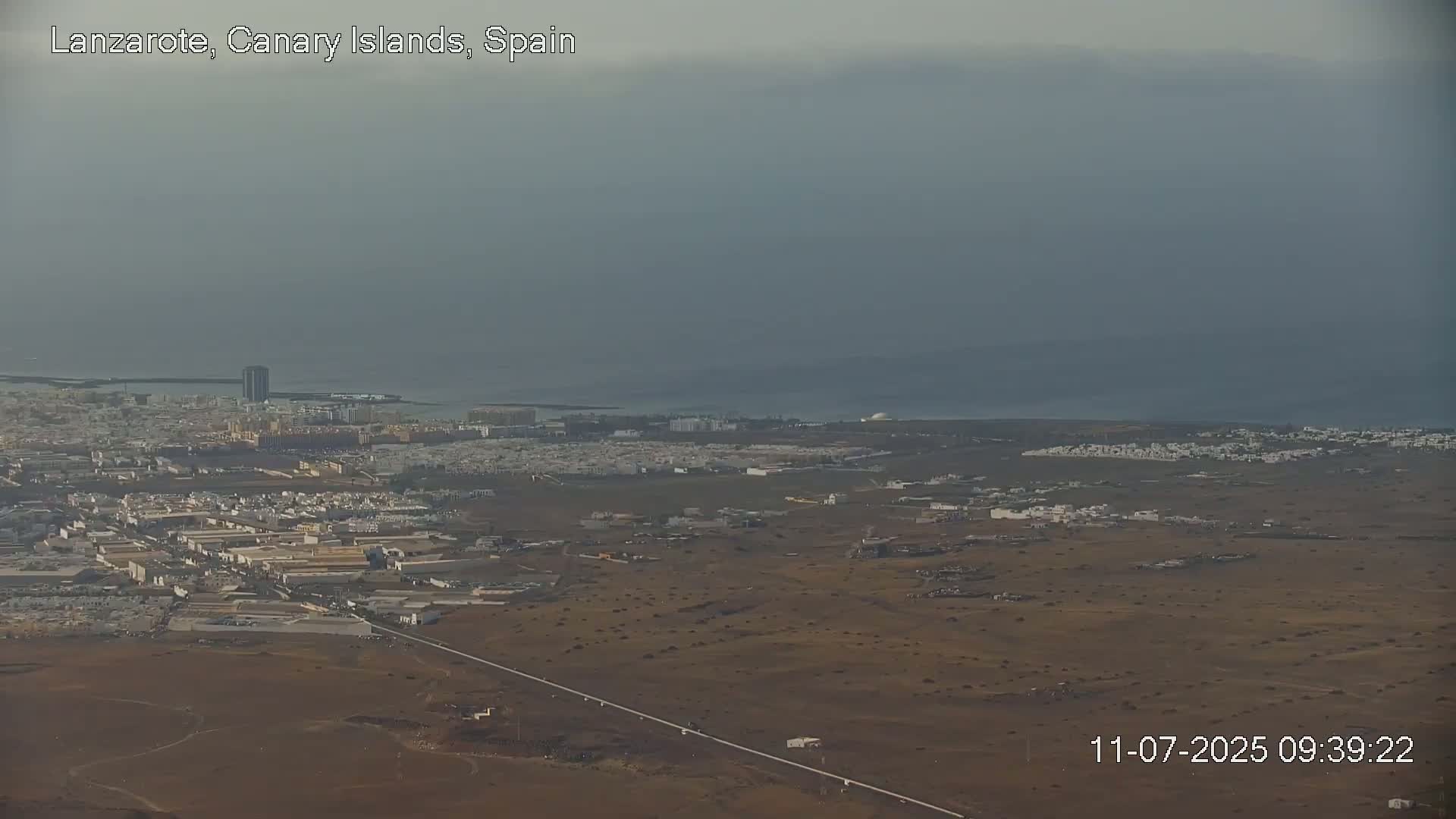 A hazy, overcast day reveals a coastal town sprawling across a flat, brown landscape that meets a calm ocean.