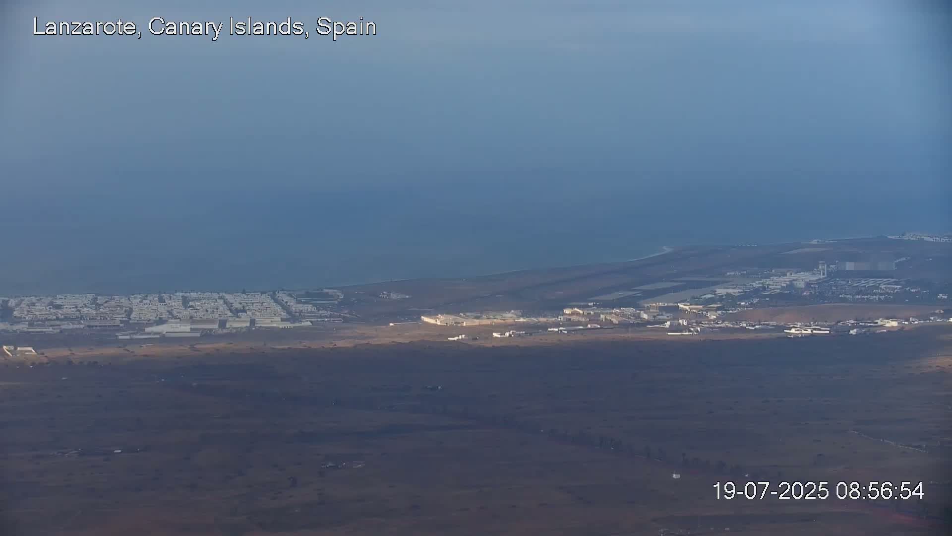 A hazy coastal view shows an airport and town near a dark blue ocean under a mostly clear sky.