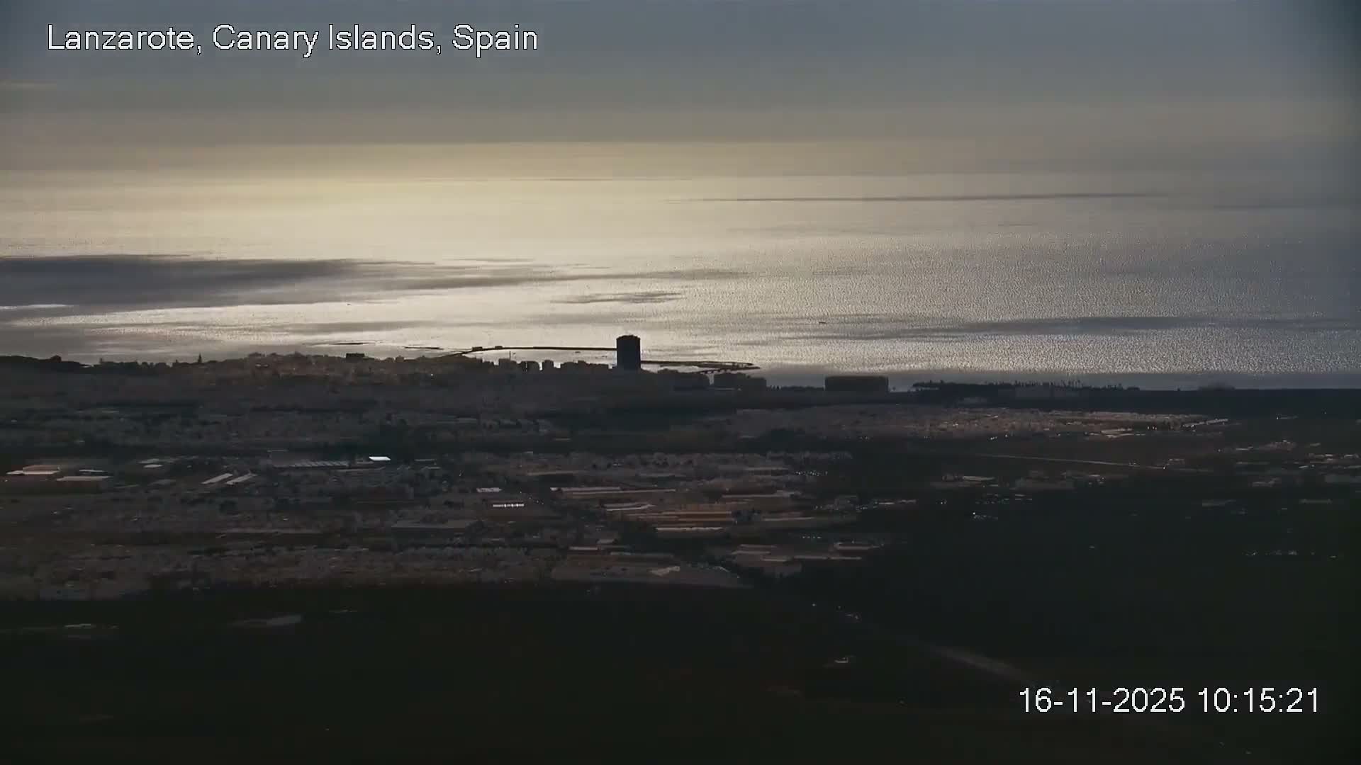 An elevated panoramic view reveals a coastal city with a prominent dark tower stretching along a shimmering ocean under a partly cloudy sky.
