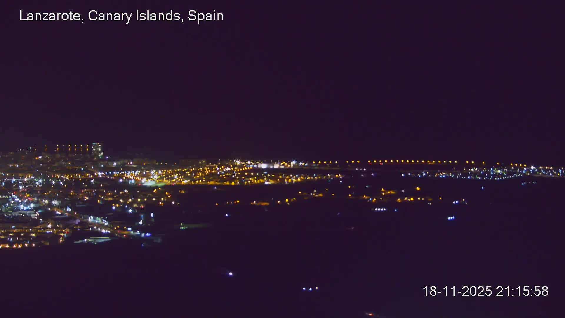 An elevated panoramic view reveals a coastal city with a prominent dark tower stretching along a shimmering ocean under a partly cloudy sky.