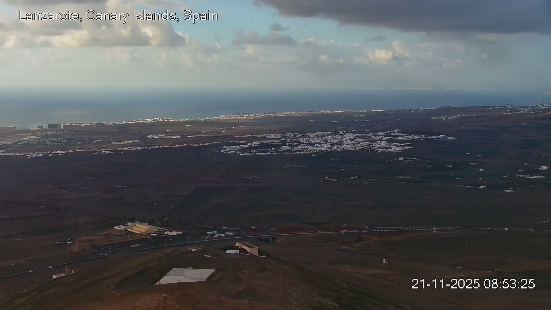 An expansive aerial view reveals a vast coastal landscape with dark, arid terrain dotted by numerous white-built towns, stretching towards a deep blue sea under a partly cloudy sky.