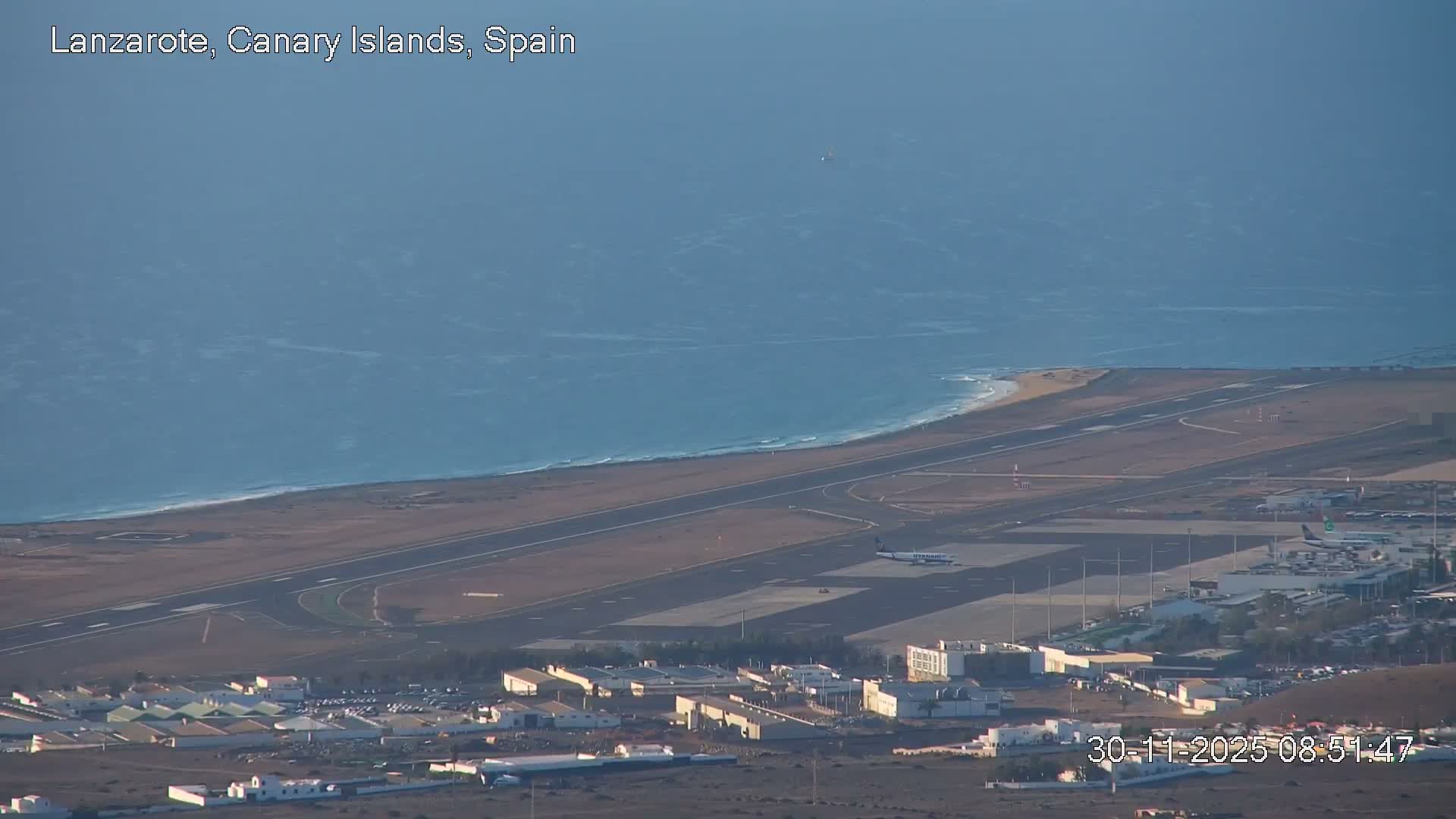 A bright, sunny aerial view shows an airport bustling with planes near terminal buildings, situated on a sandy coastline beside a blue, choppy ocean under a clear sky.