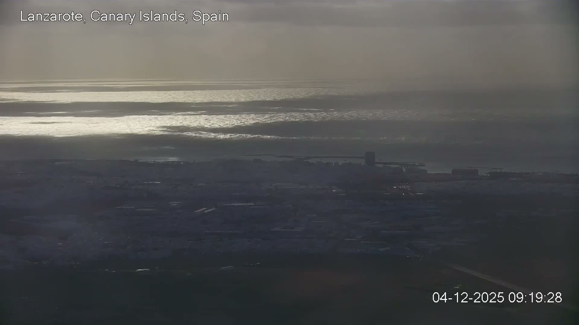An elevated view reveals a hazy, overcast coastal city stretching towards a calm body of water with streaks of bright light reflecting on its surface under a cloudy morning sky.