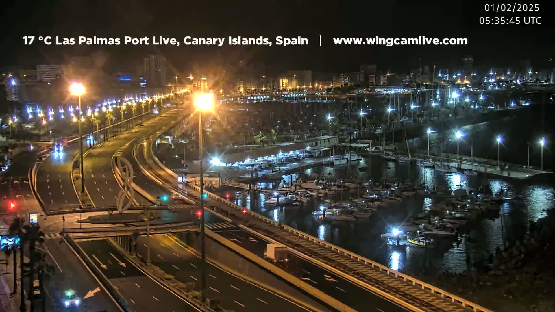 A nighttime, brightly lit view of a marina filled with boats, next to a multi-lane highway with sparse traffic.