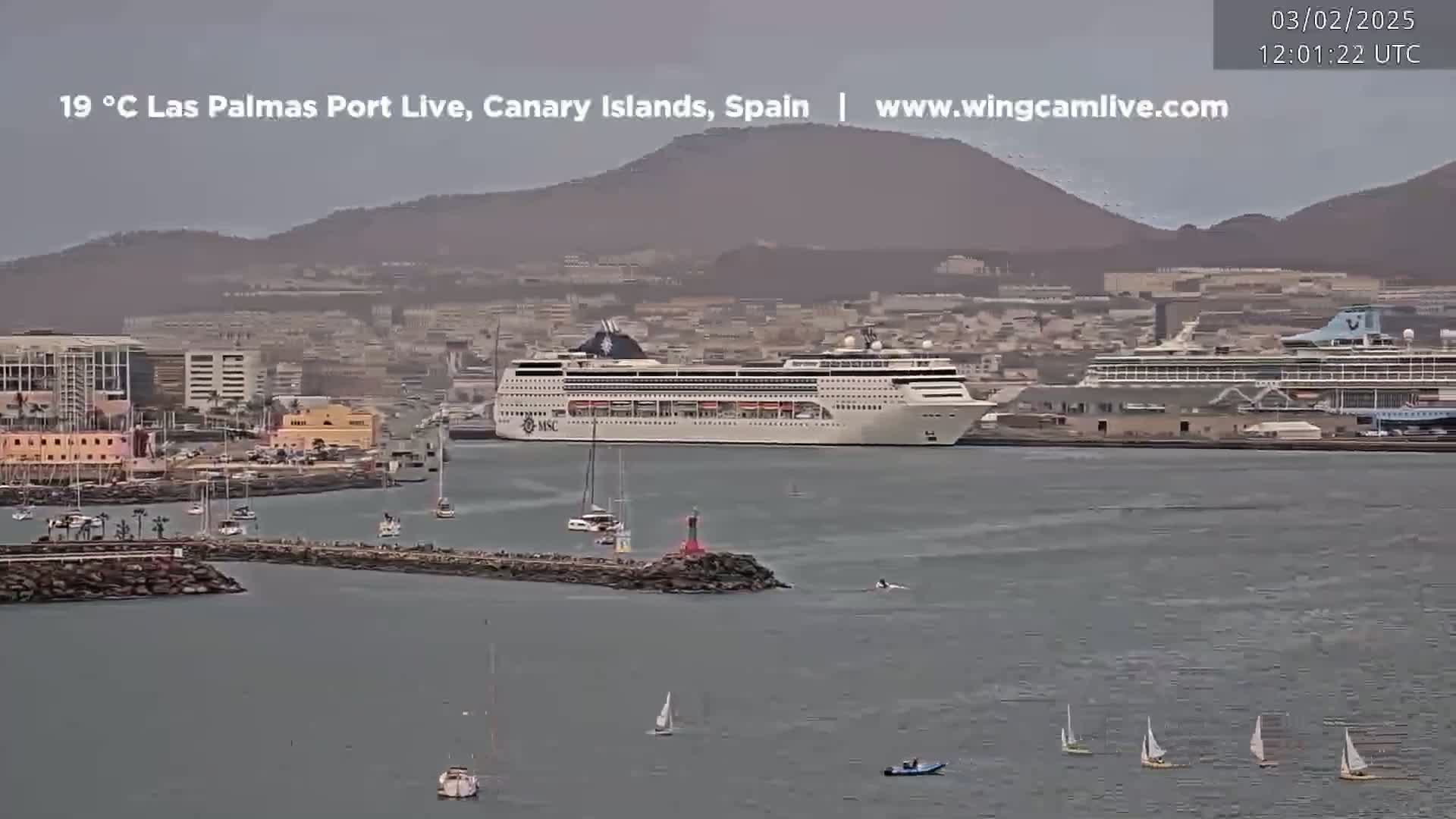 A large cruise ship is docked in a harbor, with smaller sailboats and other vessels in the water, under an overcast sky.