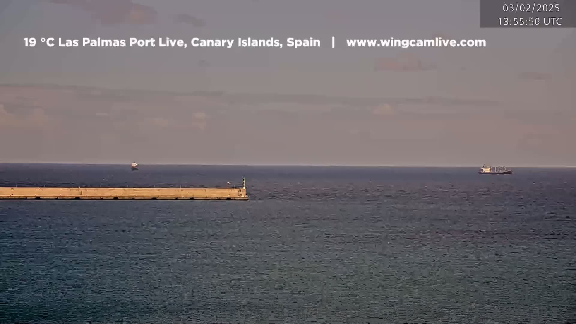 A calm ocean scene shows a long breakwater with a lighthouse at the end, a few ships in the distance, and a mostly clear sky.