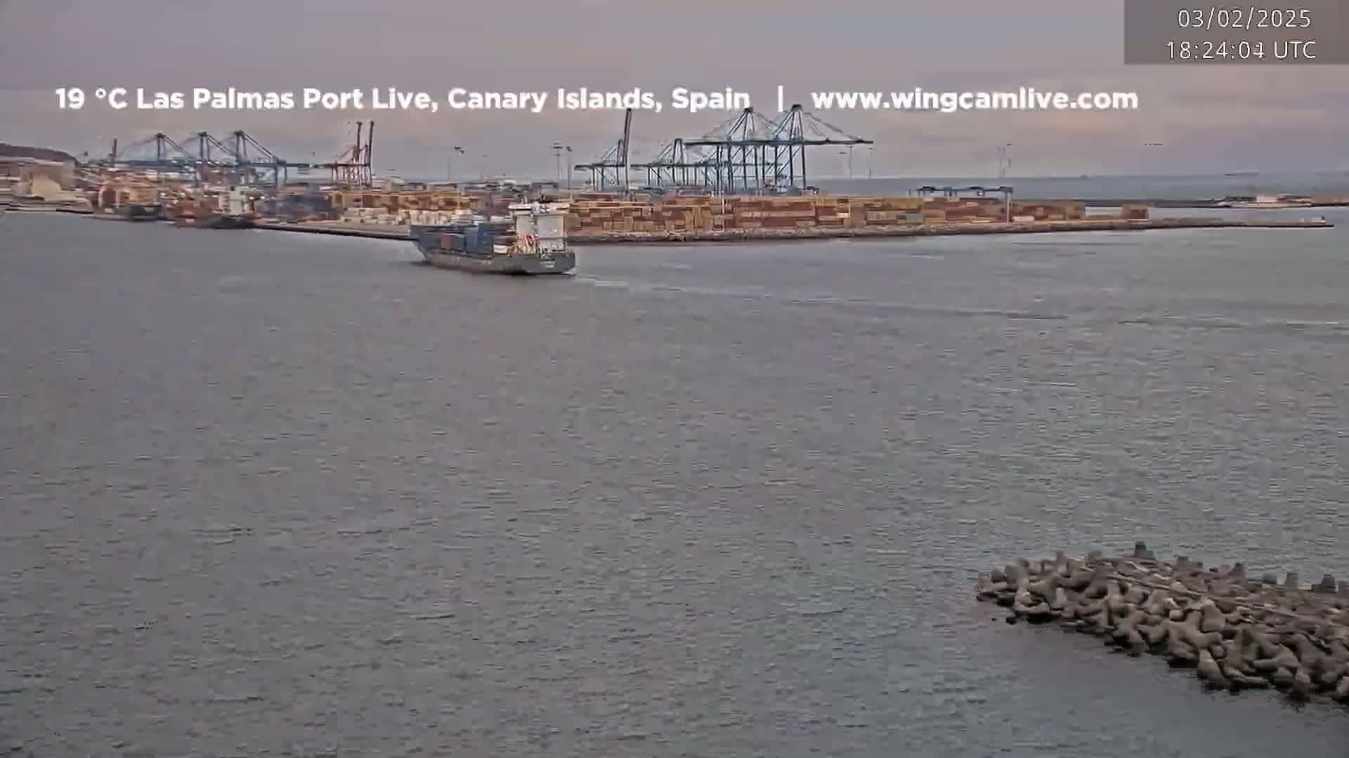 A container ship sails past a large port with several cranes under an overcast sky.