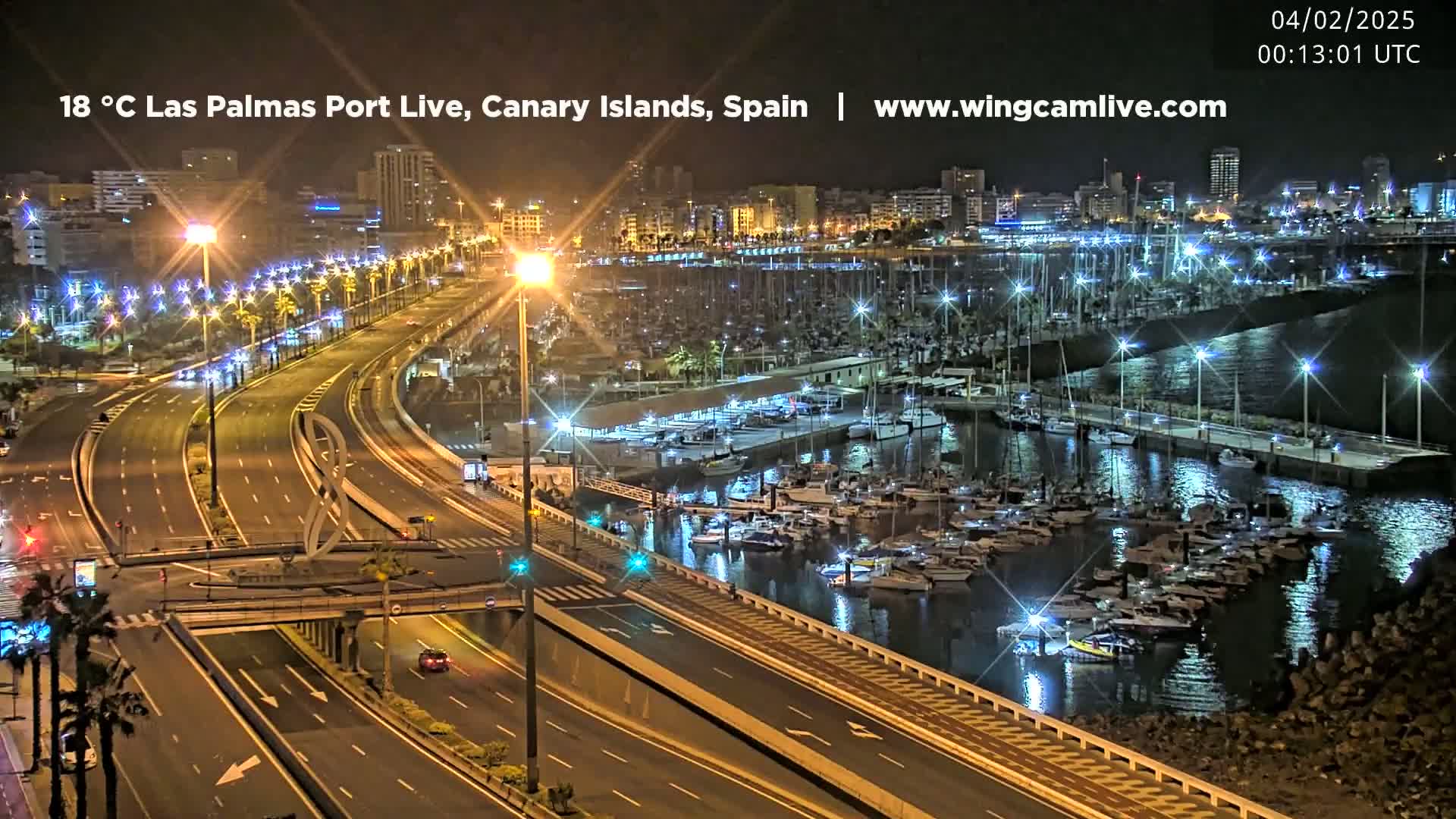A nighttime, brightly lit view of a harbor filled with boats next to a multi-lane highway and a city skyline.