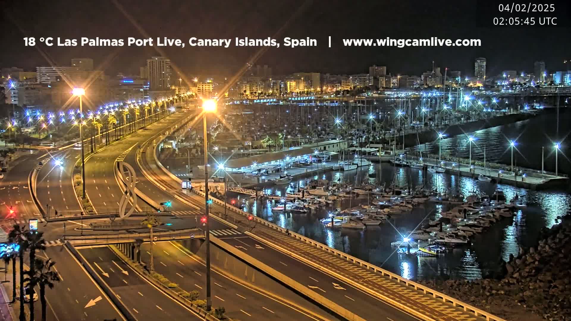 A nighttime view of a city's harbor filled with boats, next to a multi-lane highway with bright streetlights.