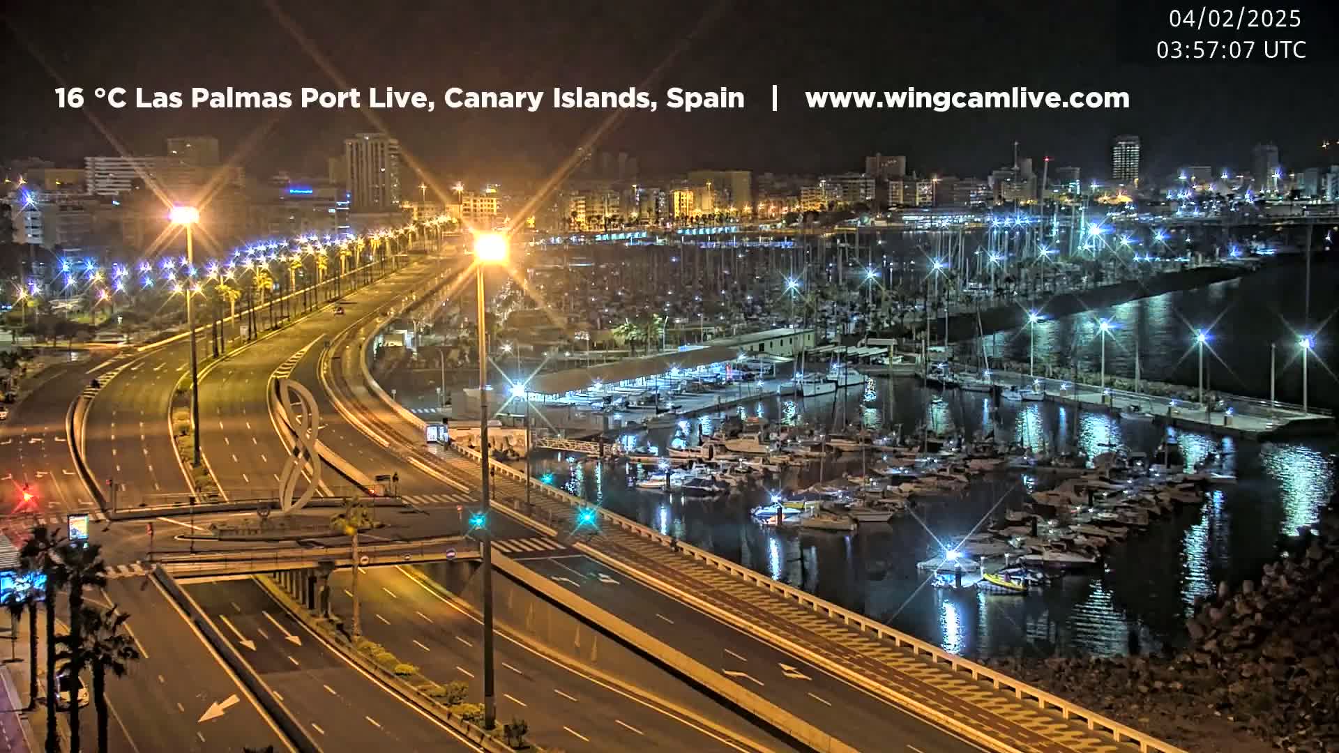 A nighttime view of a harbor filled with boats next to a multi-lane highway, with city lights illuminating the scene.