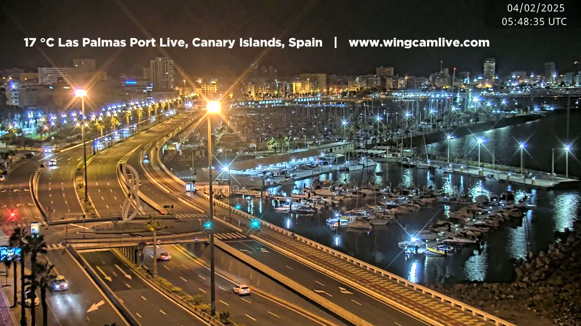 A nighttime, well-lit view of a marina filled with boats next to a multi-lane highway with sparse traffic.