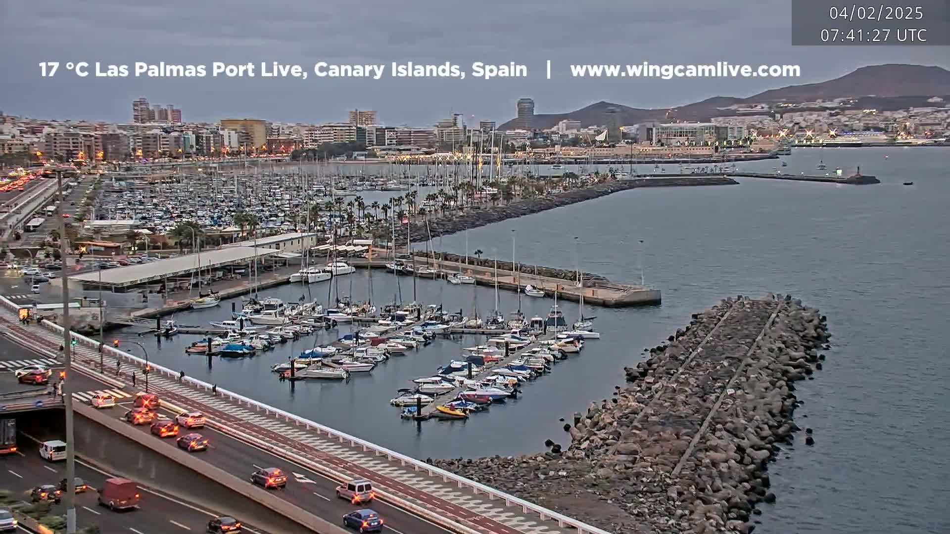 A harbor filled with numerous boats is seen from an aerial perspective, under an overcast sky, with a roadway and city buildings in the background.