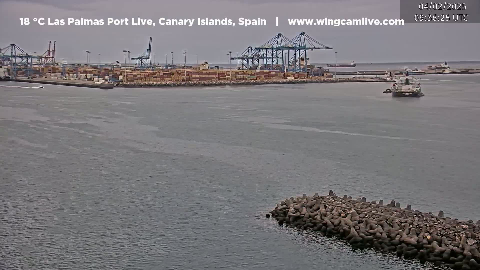 A container port with several cranes is visible across a calm body of water under an overcast sky, with a small breakwater in the foreground.