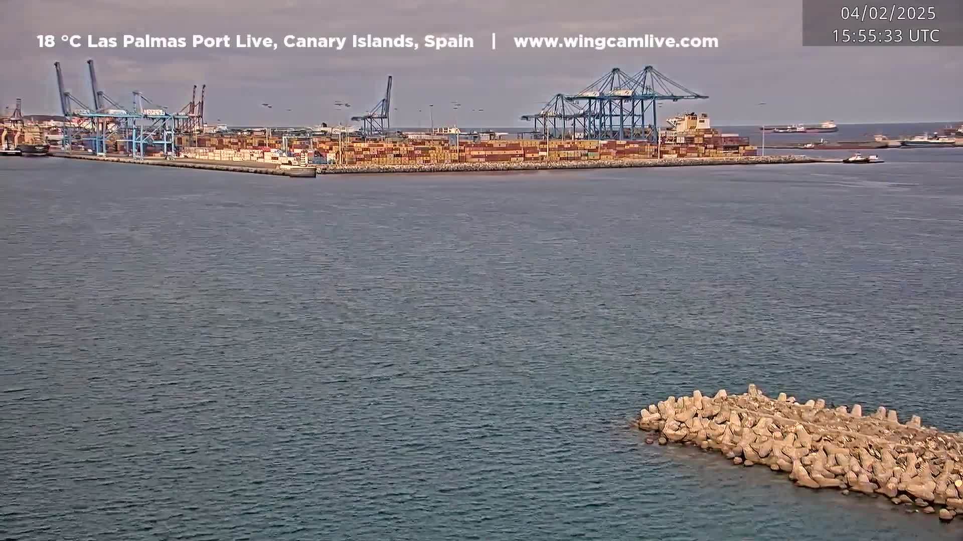 A large container port with numerous cranes and ships is visible across a calm body of water, with a small breakwater in the foreground.