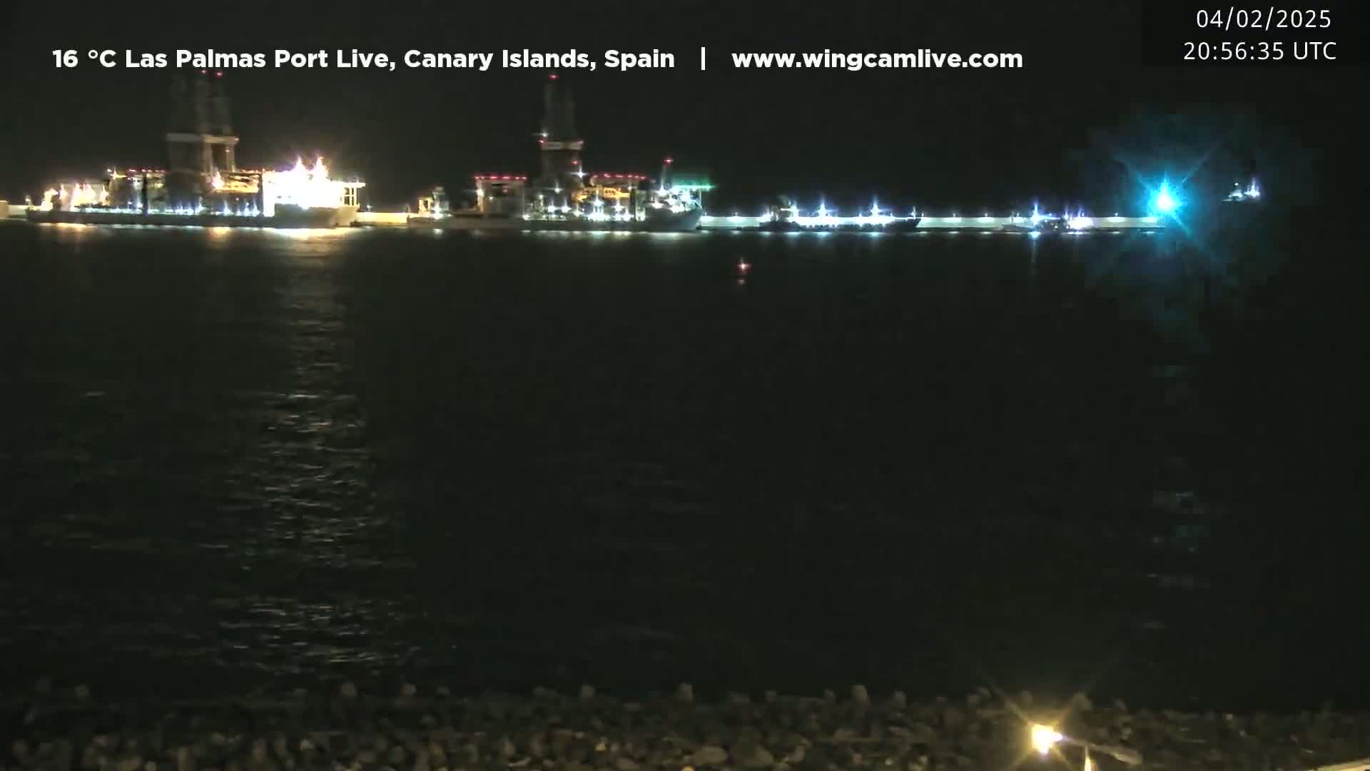 At night, brightly lit ships are moored in a calm, dark body of water, with a dark shoreline visible in the foreground.
