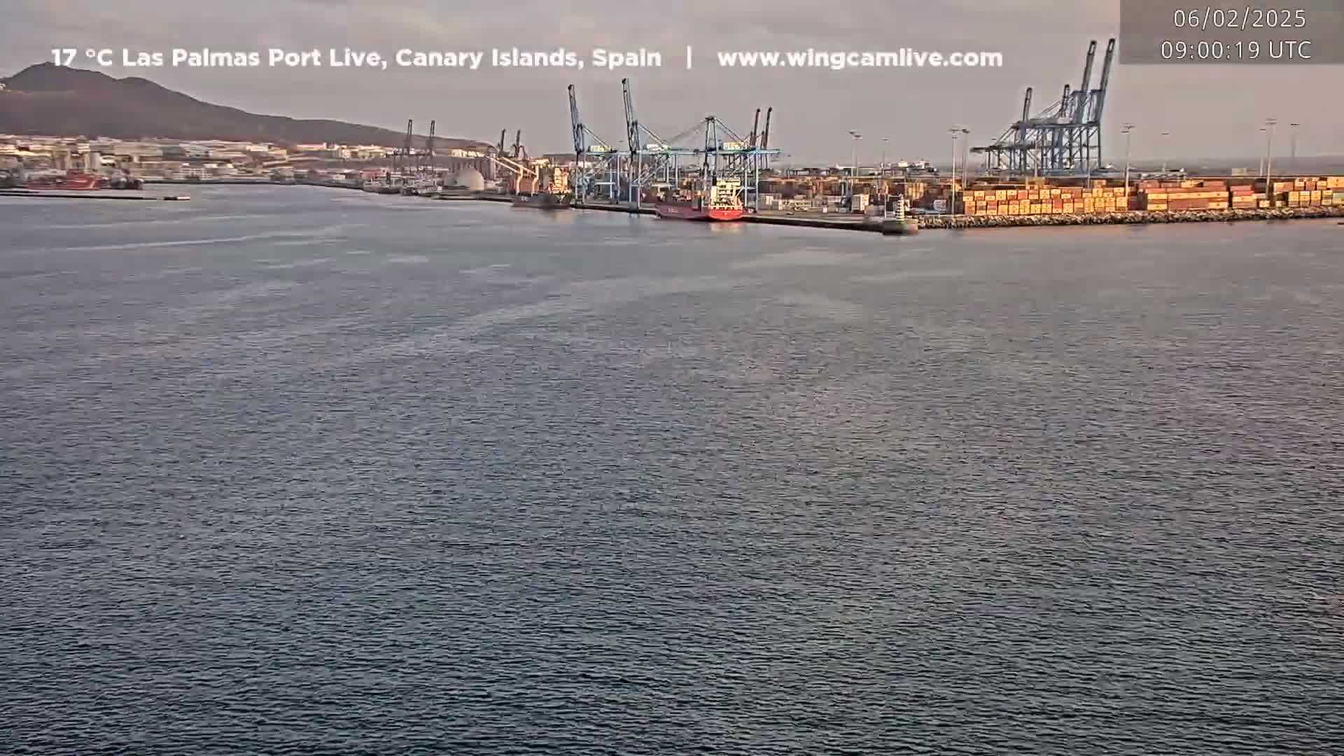 A calm body of water shows a port with several cranes, cargo ships, and containers under a mostly clear sky.