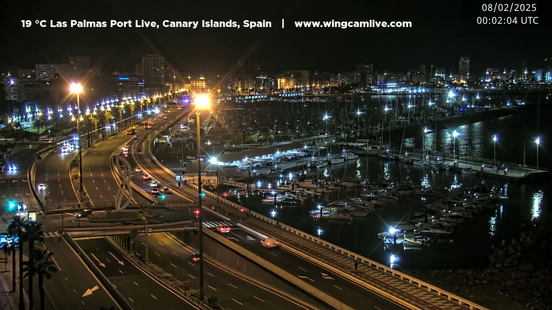 A nighttime view of a marina filled with boats, next to a brightly lit highway with cars driving on it.
