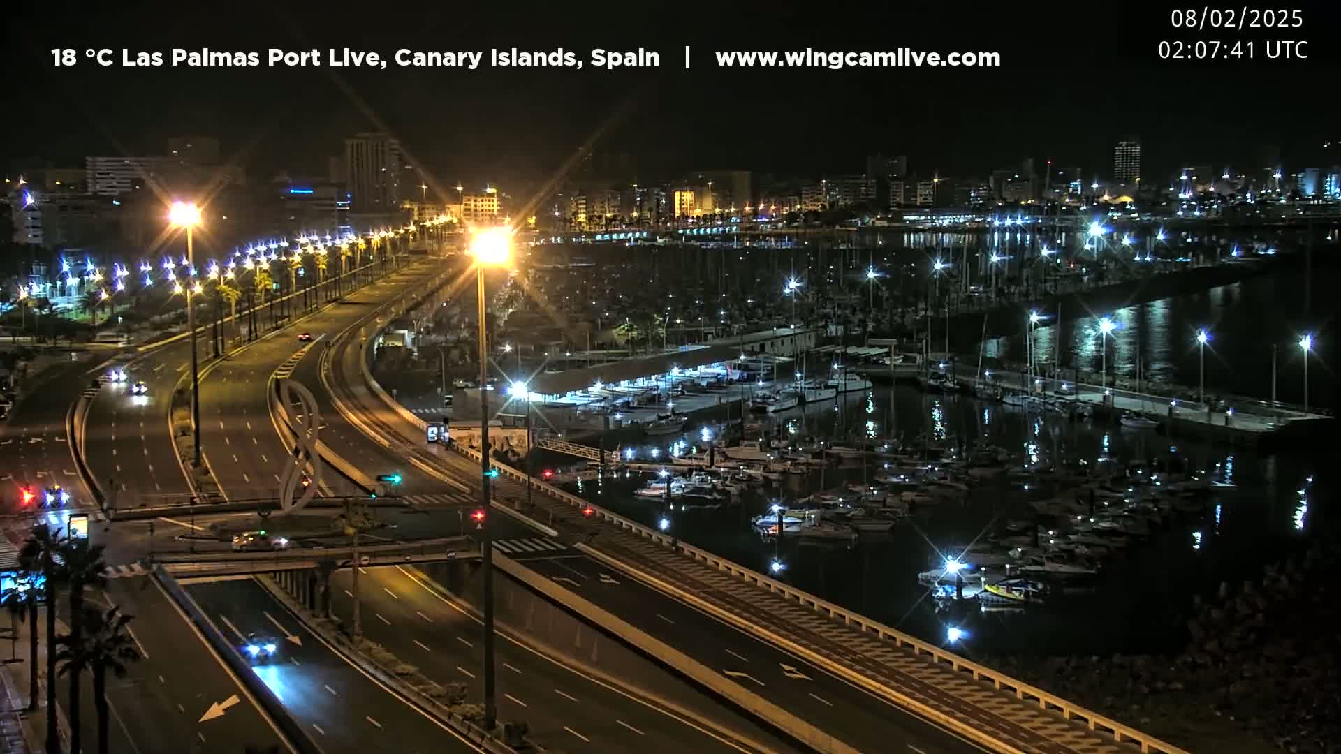 A brightly lit city harbor at night filled with numerous boats, overlooked by a multi-lane highway.