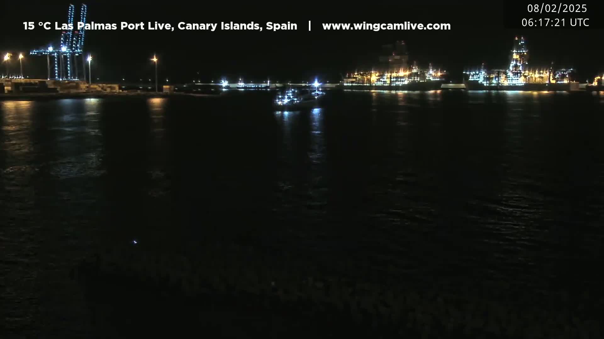 At night, a dark body of water reflects the lights of a port, with several ships and cranes visible along the shoreline.