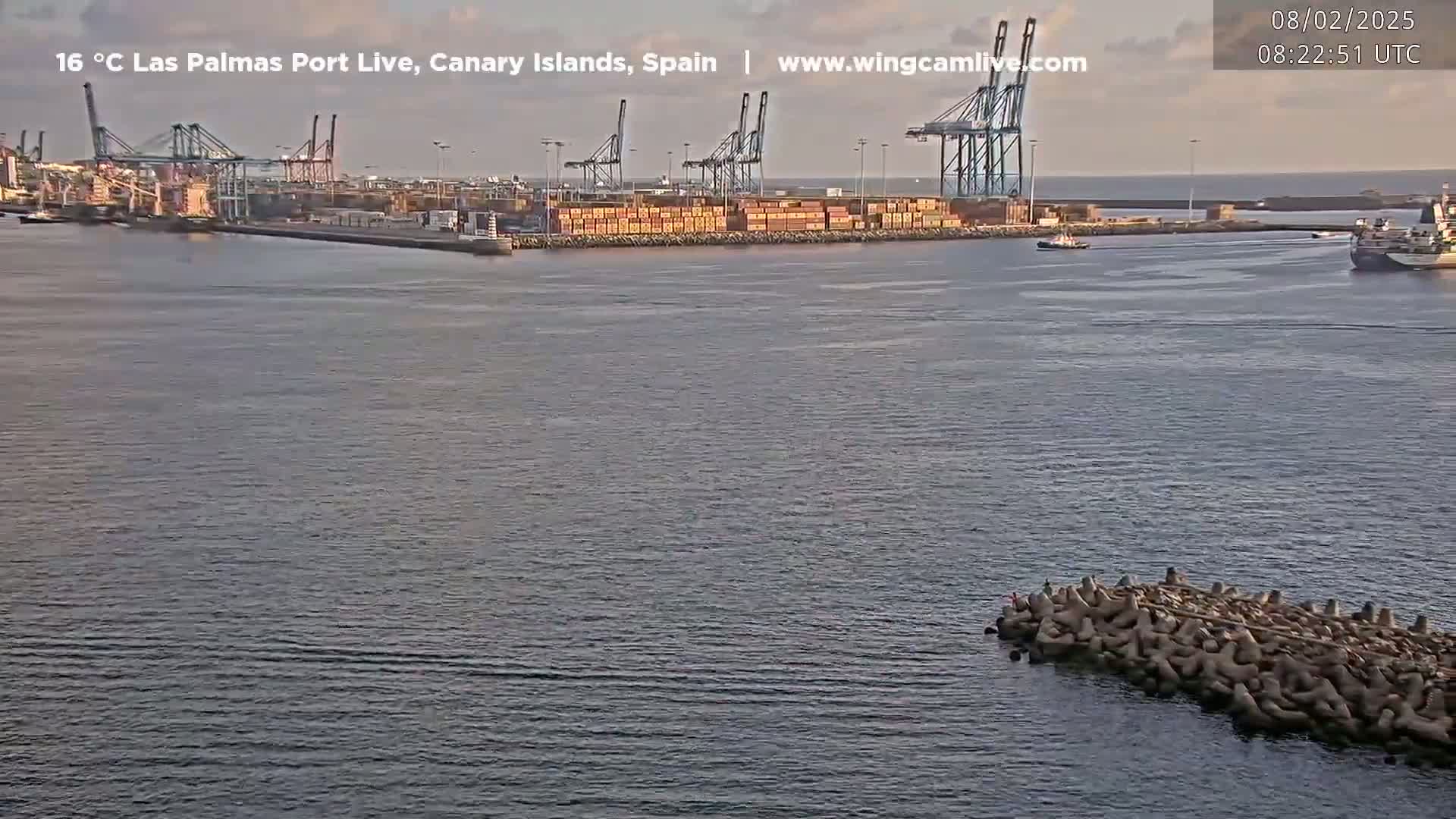 A calm harbor scene shows a container port with several cranes and ships under a mostly clear sky.