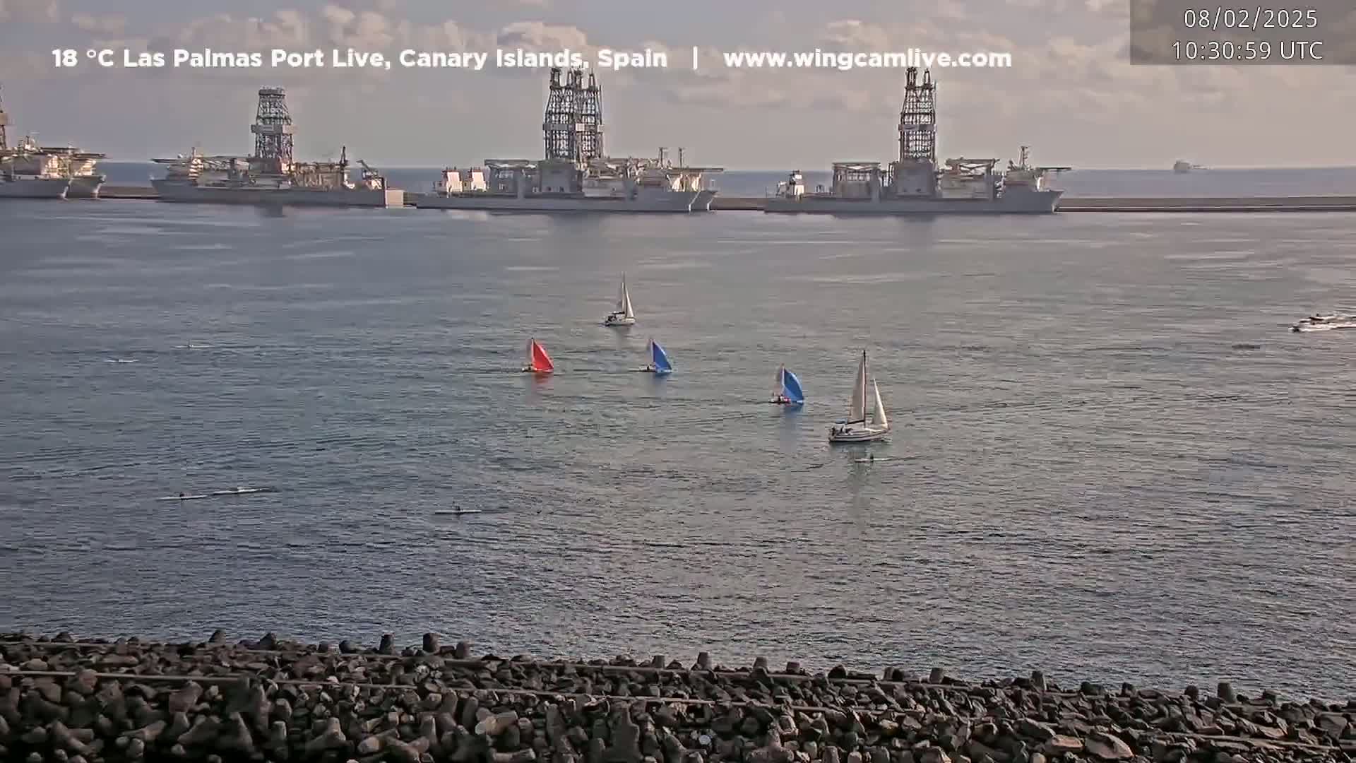 Several sailboats are on calm water in front of a port with several large ships docked under a partly cloudy sky.