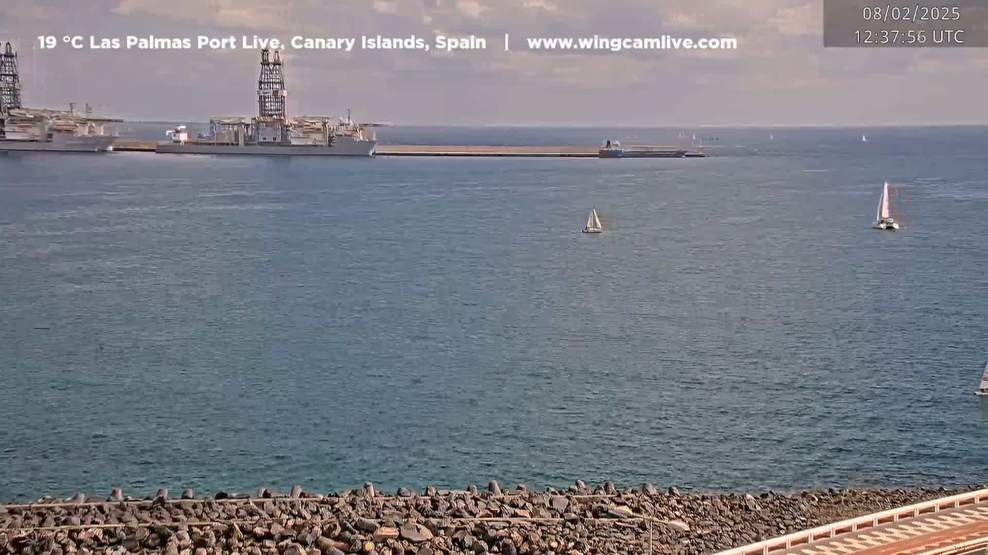 A mostly sunny day shows a calm harbor with several ships at dock and a few sailboats in the distance.