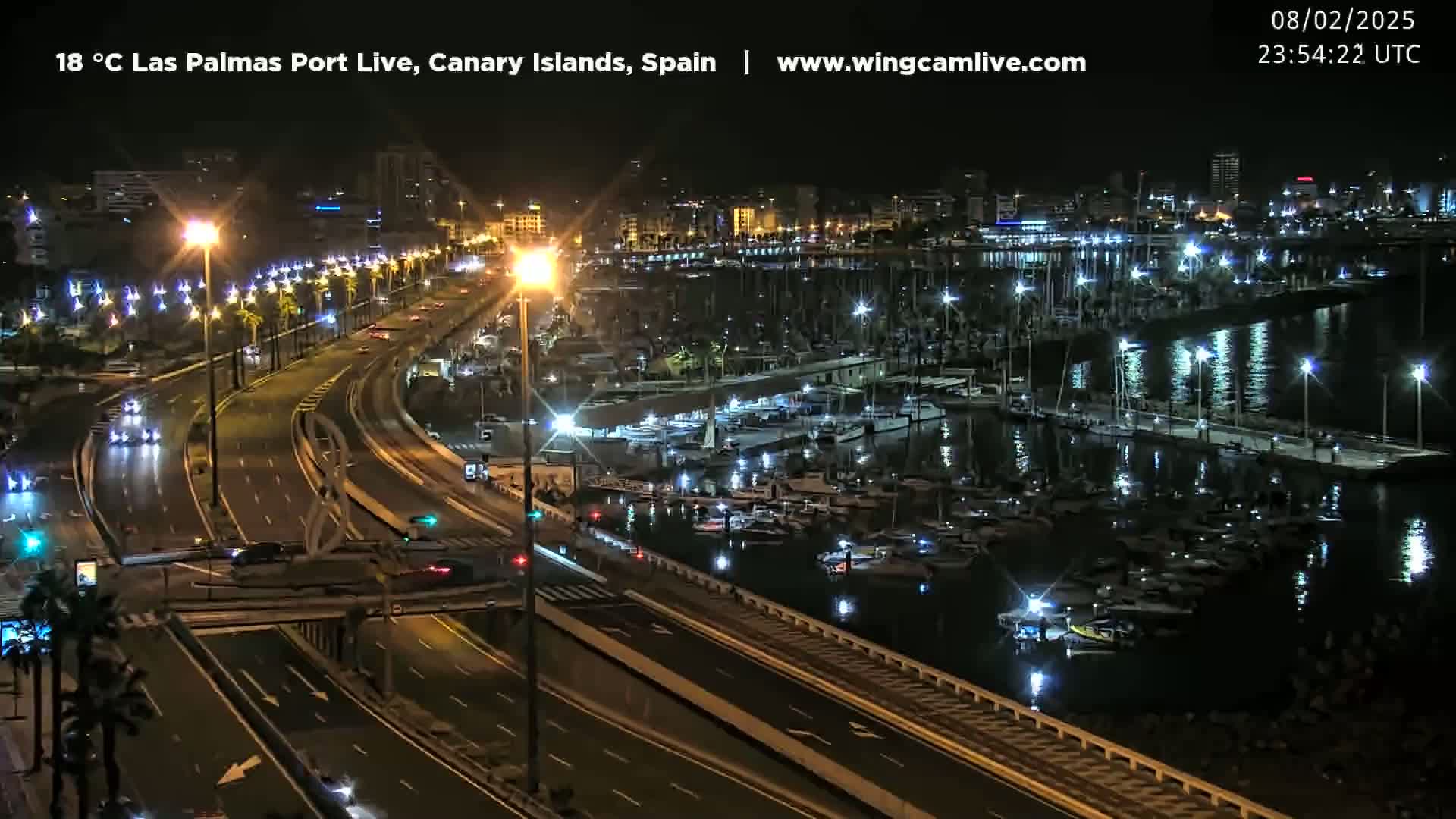 A nighttime, high-angle view of a harbor filled with boats, next to a multi-lane highway with city buildings in the background.