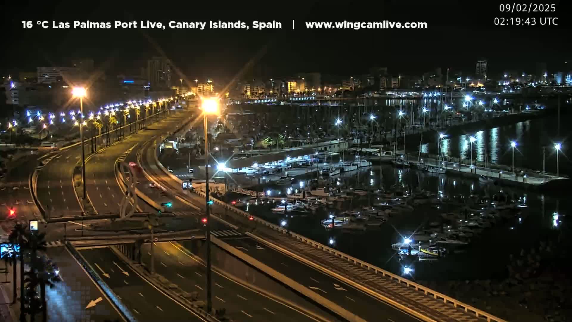 A nighttime, brightly lit highway overlooks a calm harbor filled with boats, all under a clear sky.