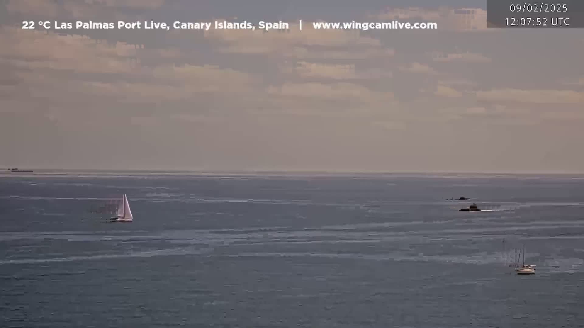 A calm, slightly cloudy day shows a wide expanse of ocean with several small sailboats and a dark vessel in the distance.
