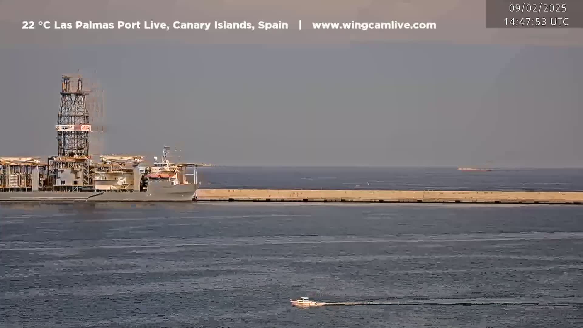 A large drilling ship is docked near a long breakwater in calm, hazy conditions.