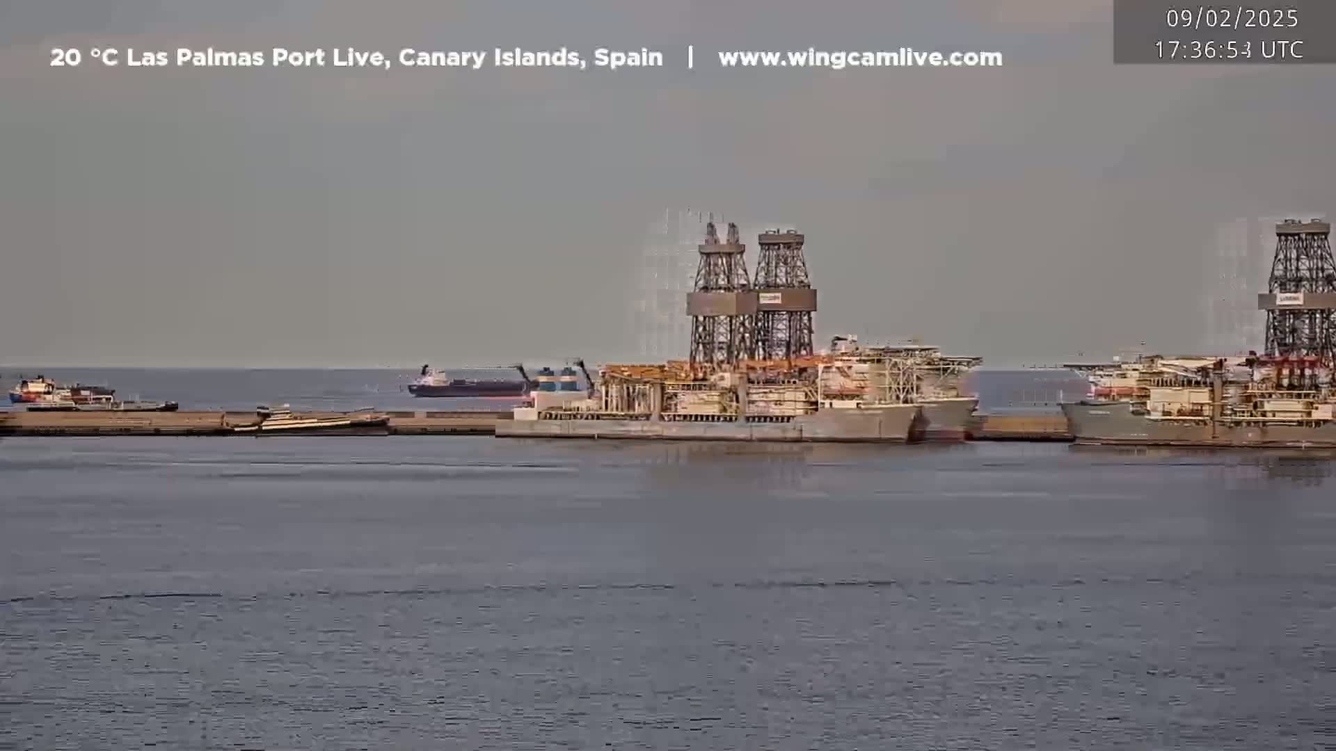 Several large ships, including at least one drillship, are docked at a port under an overcast sky.