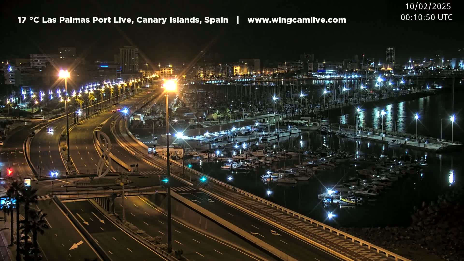 A nighttime, brightly lit city harbor filled with boats and calm water is bordered by a multi-lane highway.
