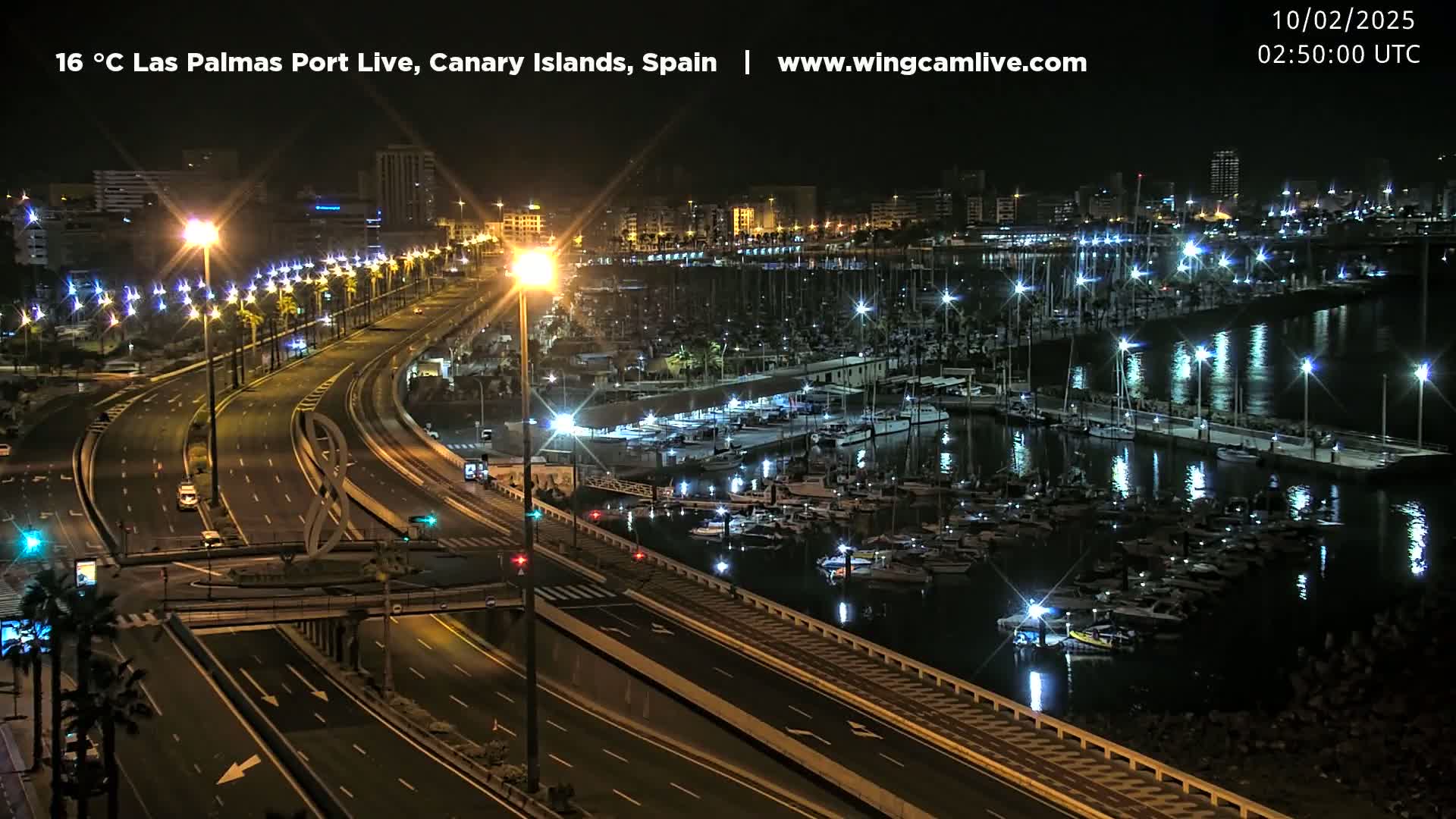 A nighttime, aerial view of a harbor filled with boats and yachts, next to a multi-lane highway with minimal traffic, under clear skies.