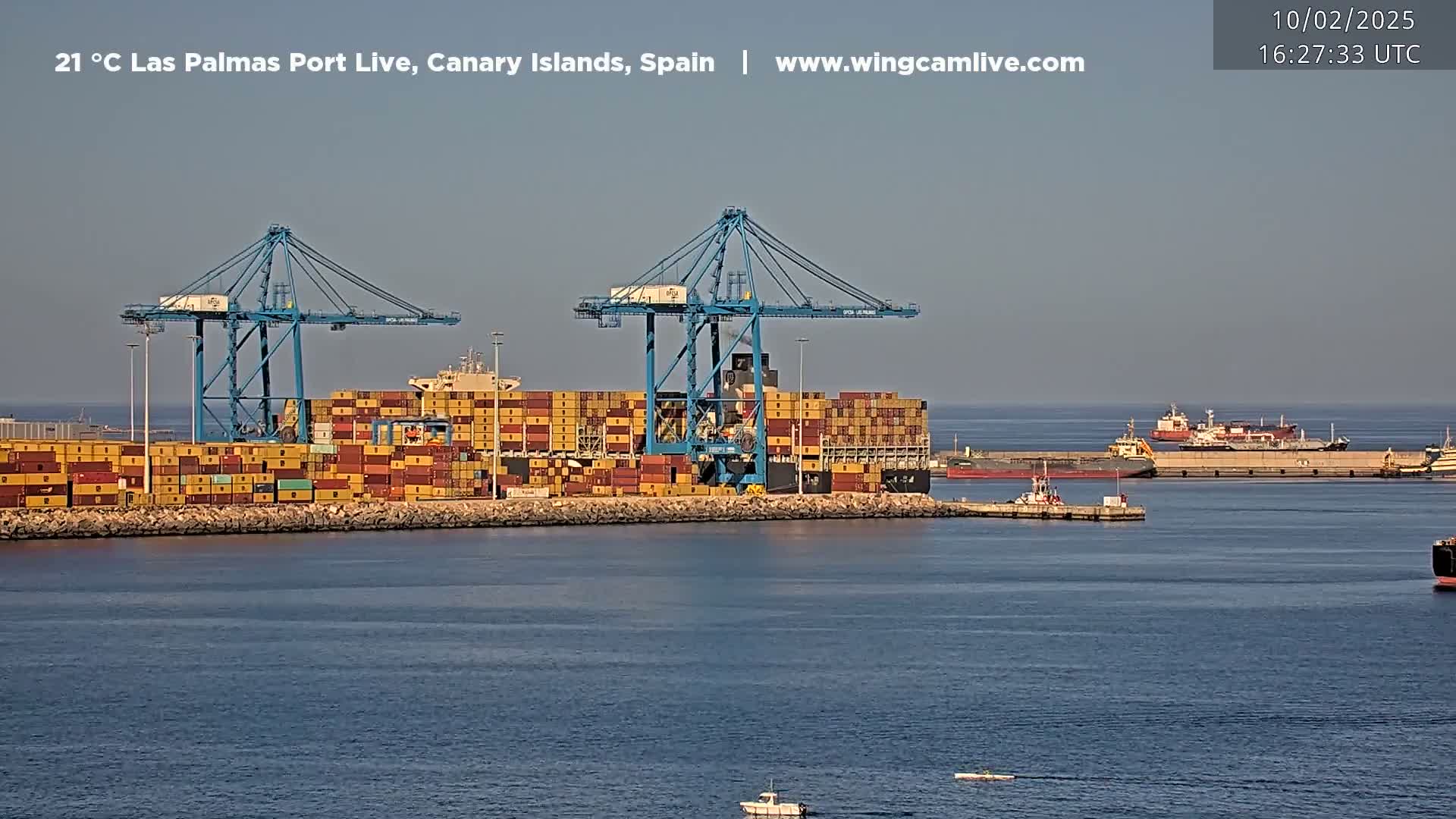 A container ship is docked at a port under a clear sky, with other vessels in the harbor.