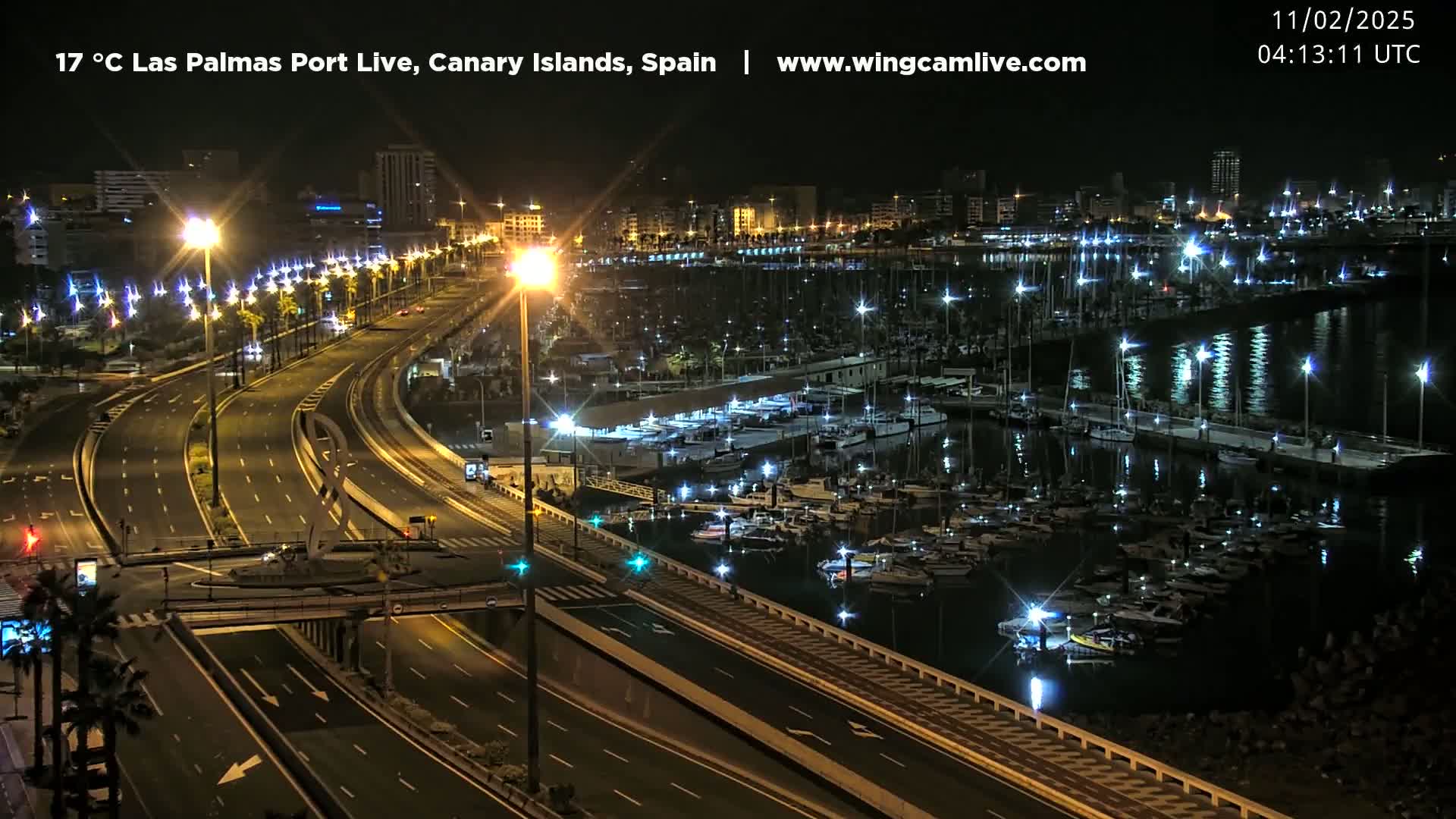 A nighttime view of a harbor filled with boats, next to a multi-lane highway, with city lights in the background.