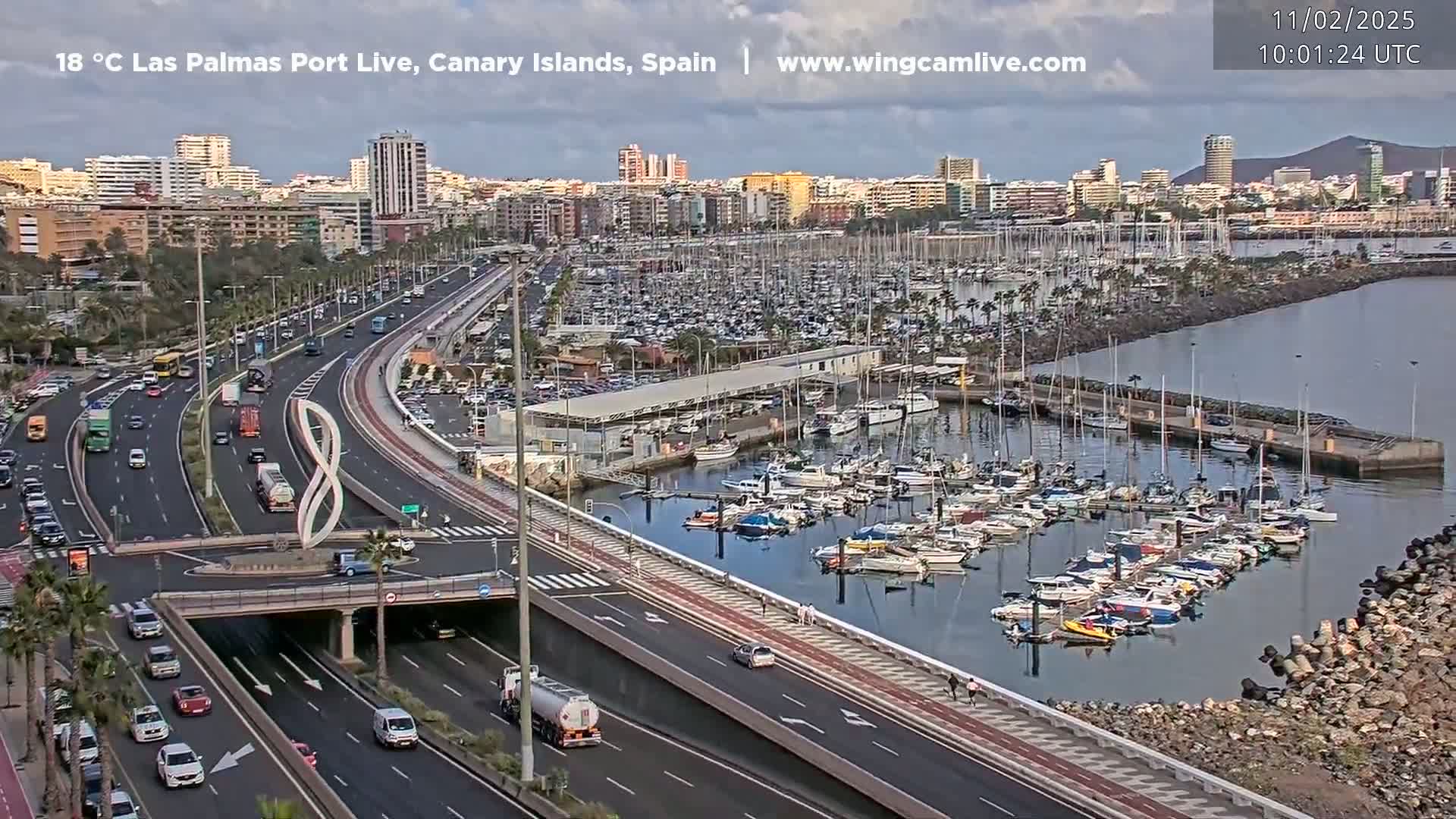 A partly cloudy day reveals a bustling harbor filled with numerous boats, adjacent to a highway with moderate traffic, all set against a backdrop of a city skyline.