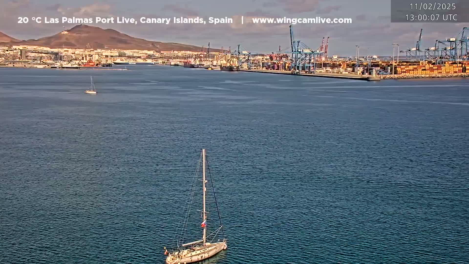 A sailboat sits in calm water in front of a port filled with cargo containers and cranes under a mostly clear sky.