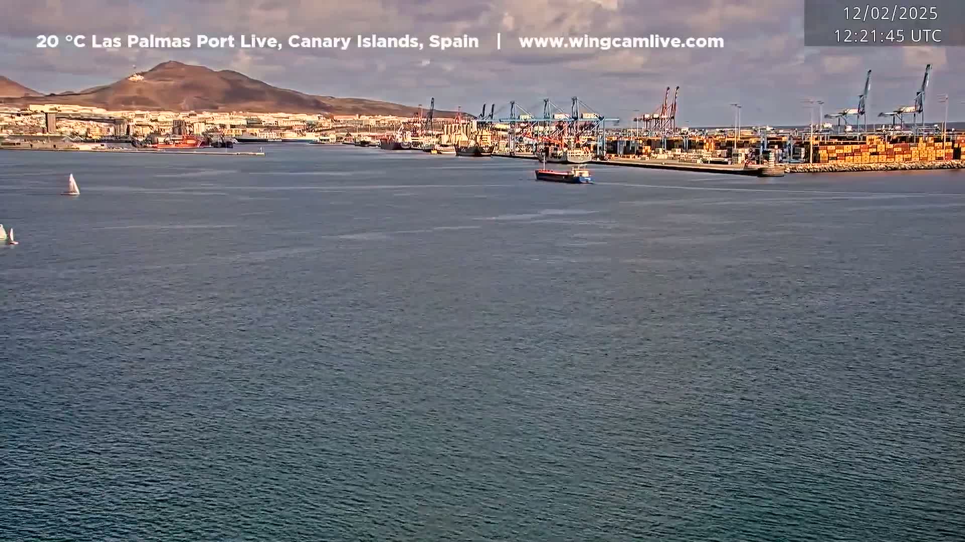 A calm body of water shows a port with several ships and containers, a city in the background, and two sailboats in the foreground under a partly cloudy sky.