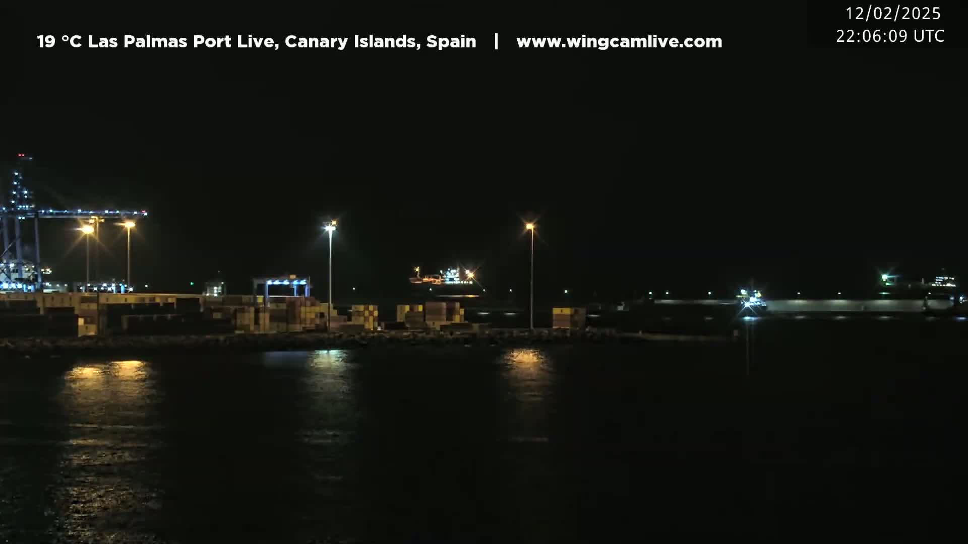 A nighttime view of a lit-up harbor with cargo containers and cranes, under a clear, dark sky.