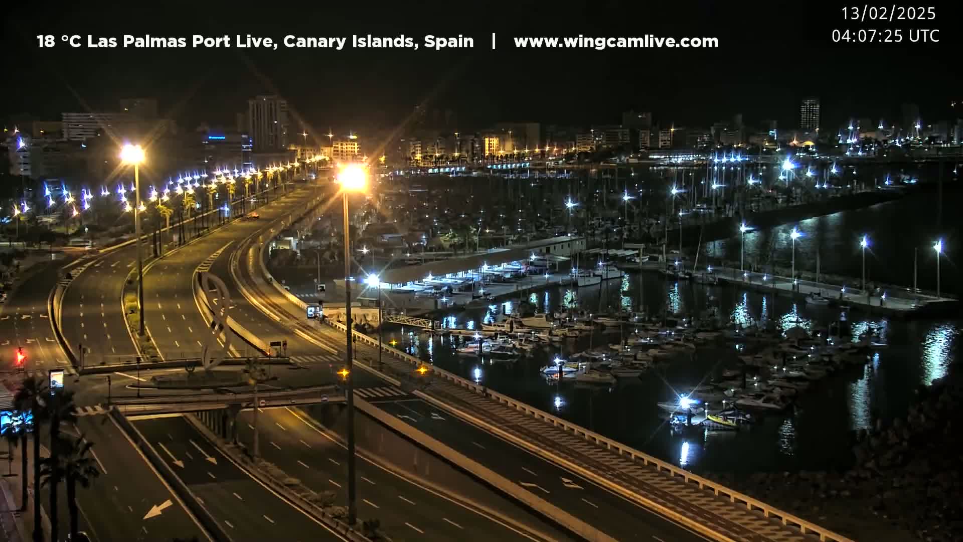 A nighttime view of a harbor filled with boats next to a multi-lane highway, with city lights in the background.