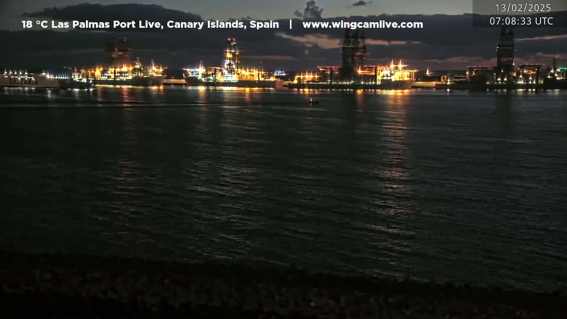 A nighttime view of a harbor filled with brightly lit ships, calm water reflecting the lights, and a dark shoreline.