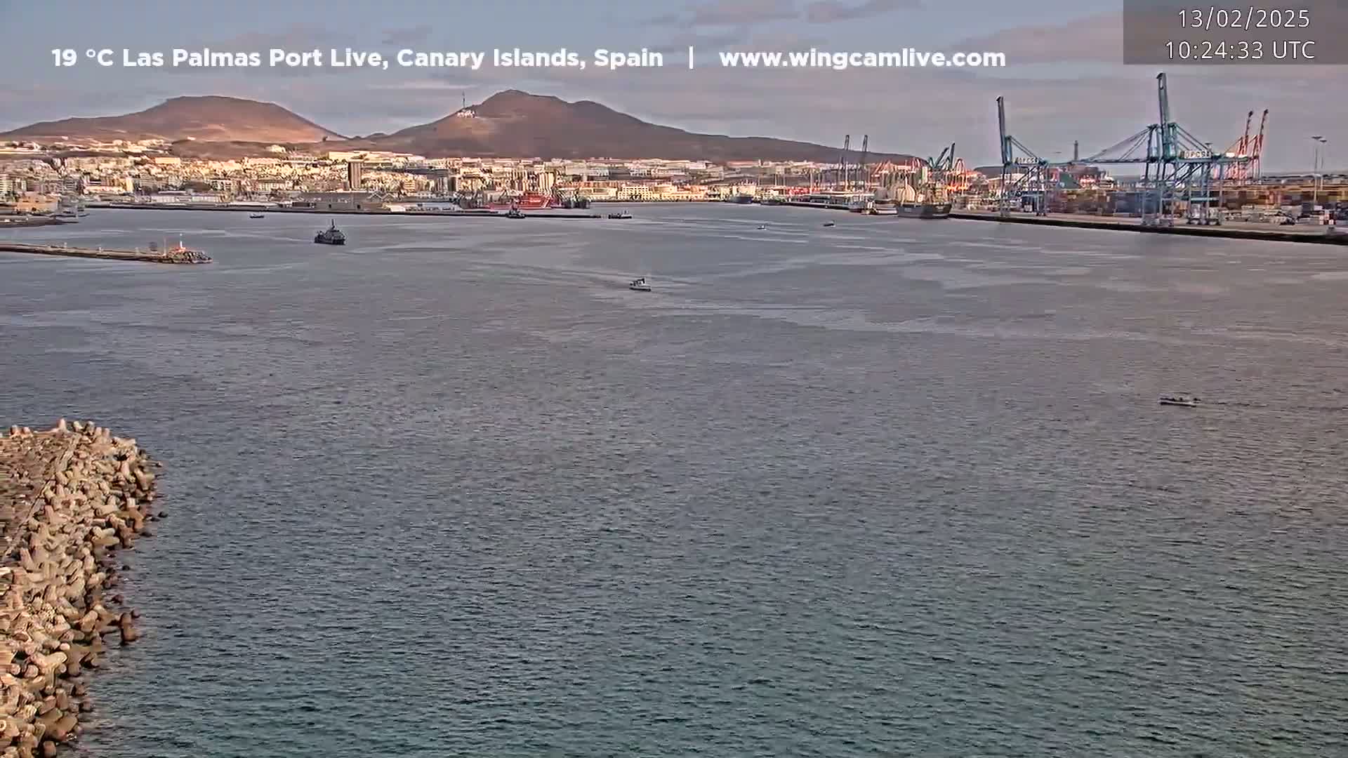 A calm harbor scene shows a city, mountains, and a busy port with several ships and cranes under a mostly clear sky.