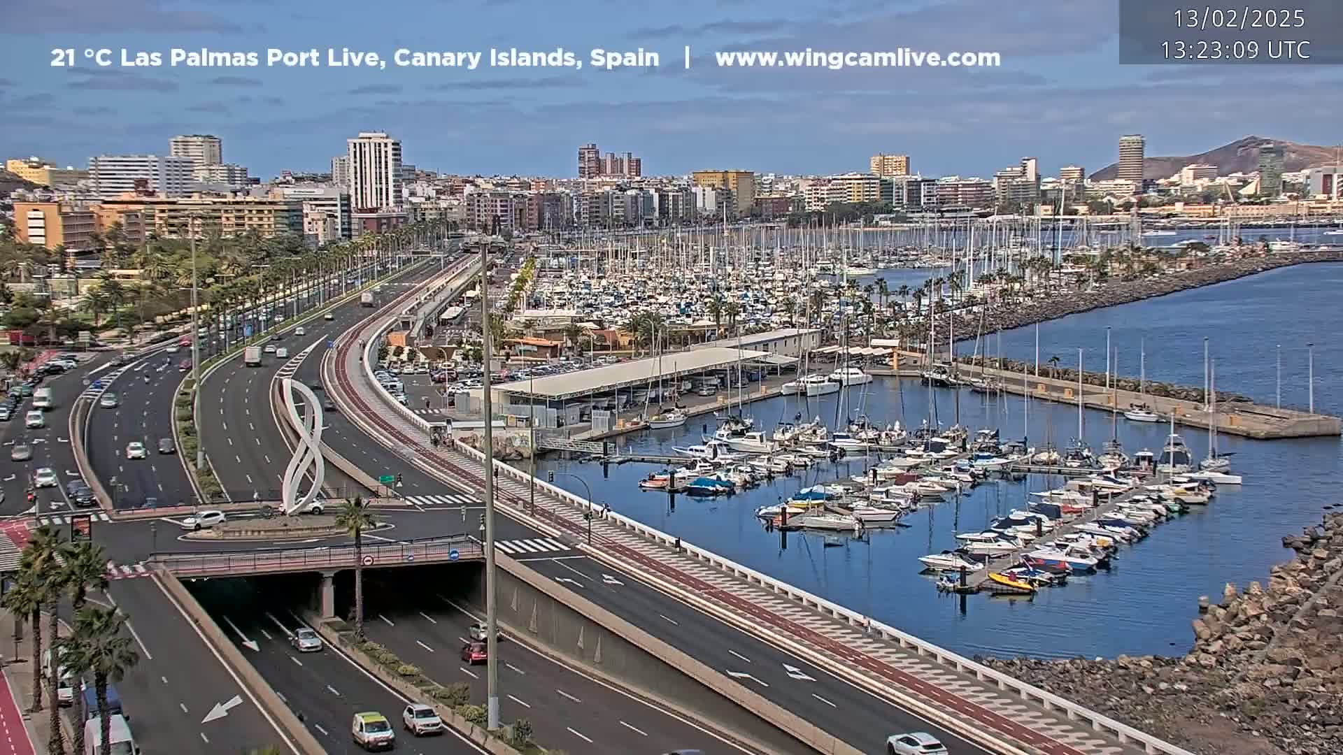 A sunny day shows a coastal city with a busy highway curving around a harbor filled with numerous boats and yachts.