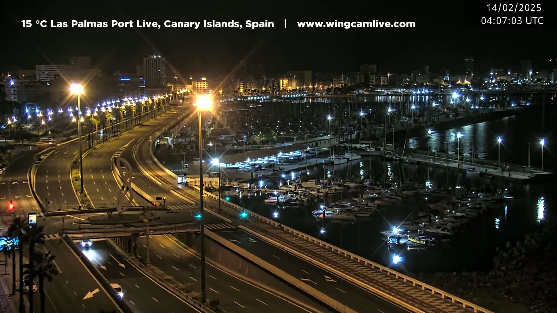 A nighttime, aerial view shows a harbor filled with boats, a city skyline, and a well-lit highway with minimal traffic.