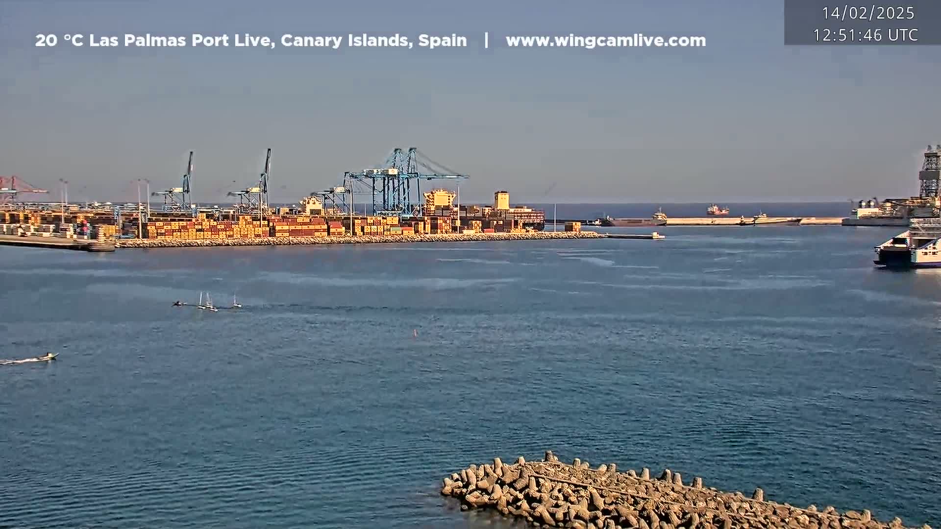 A container ship is docked at a busy port on a calm, sunny day.